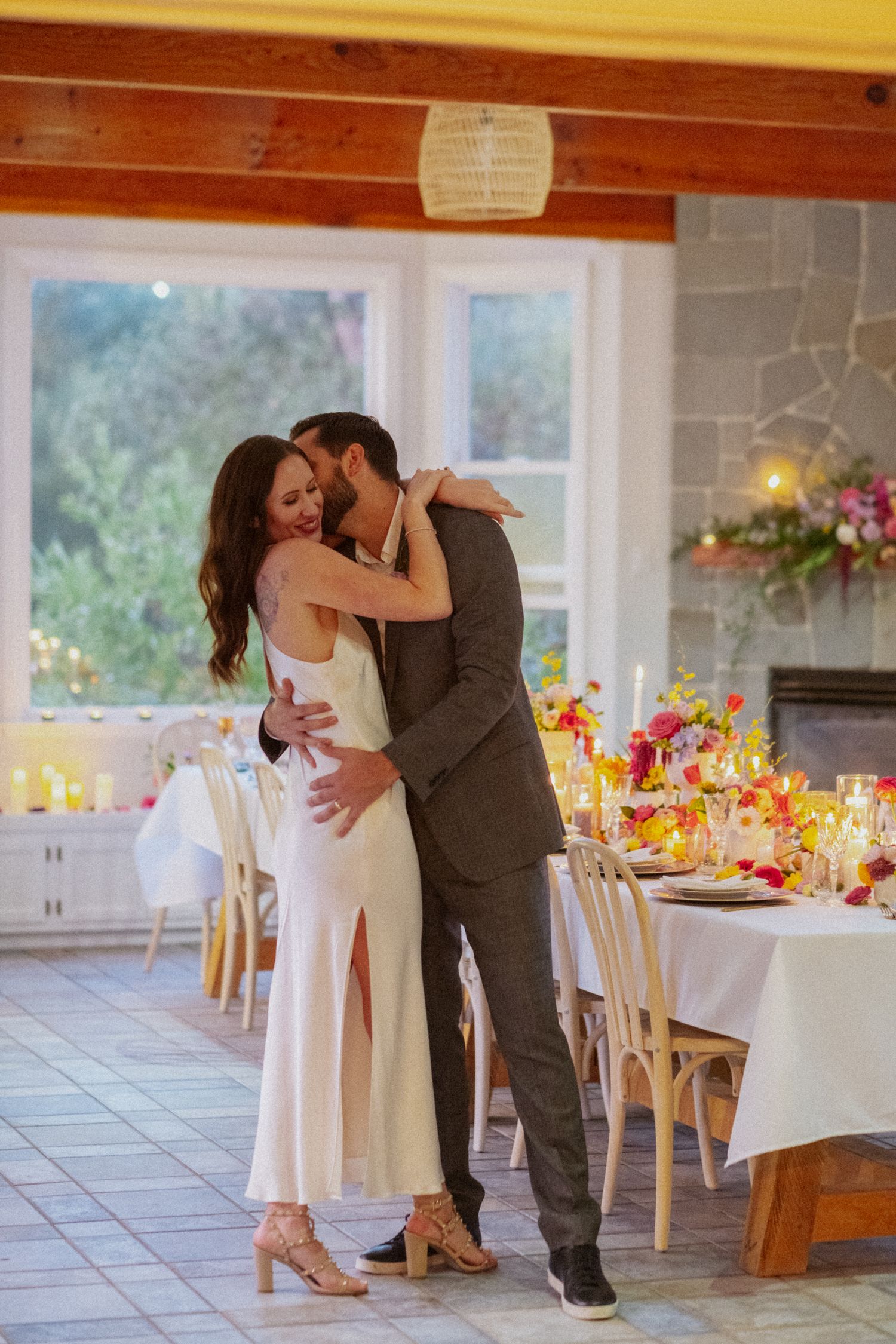 Bride and groom share romantic embrace beside candlelit reception table with vibrant floral centerpieces.