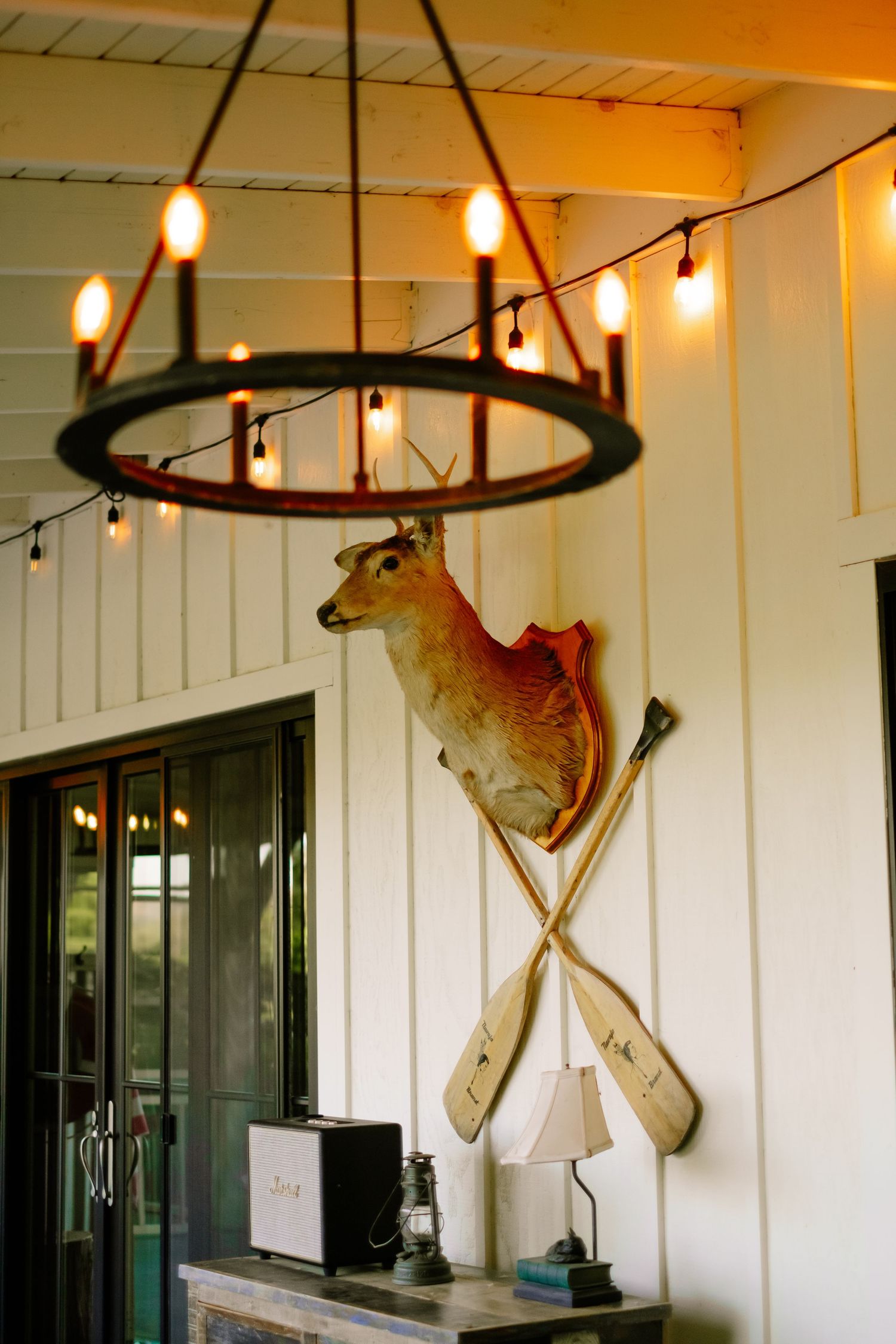 Rustic interior with wagon wheel chandelier, mounted deer head, and crossed oars on white wall.