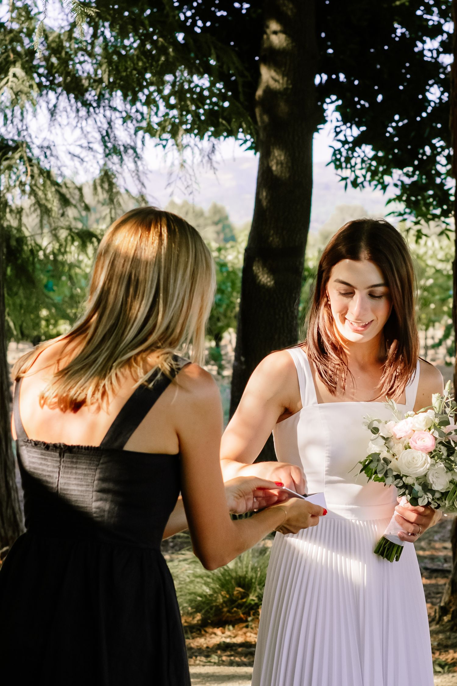 Bride in white dress holding bouquet receives document from woman in black dress outdoors.