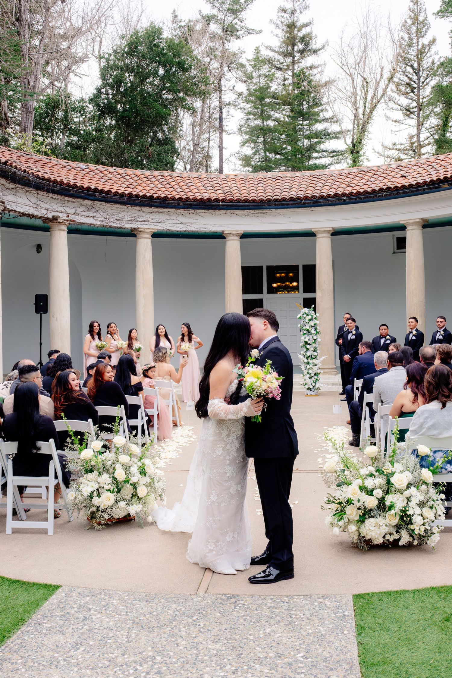Newlyweds share first kiss at outdoor wedding ceremony in elegant columned rotunda with white floral arrangements.