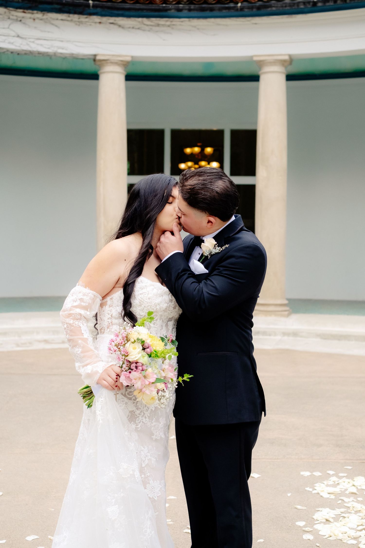 Bride and groom share a tender kiss in front of elegant columned rotunda, bride holding pink bridal bouquet.