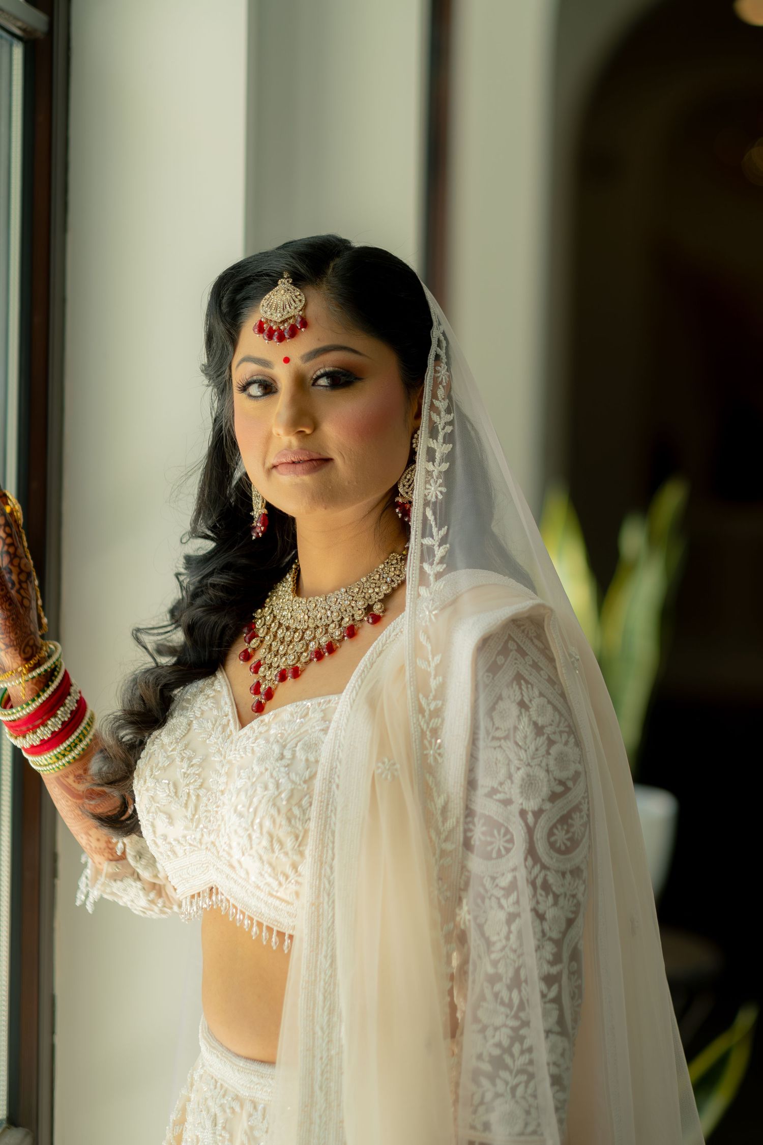 Indian bride in white lace lehenga with gold and red bridal jewelry and veil poses elegantly by a window.