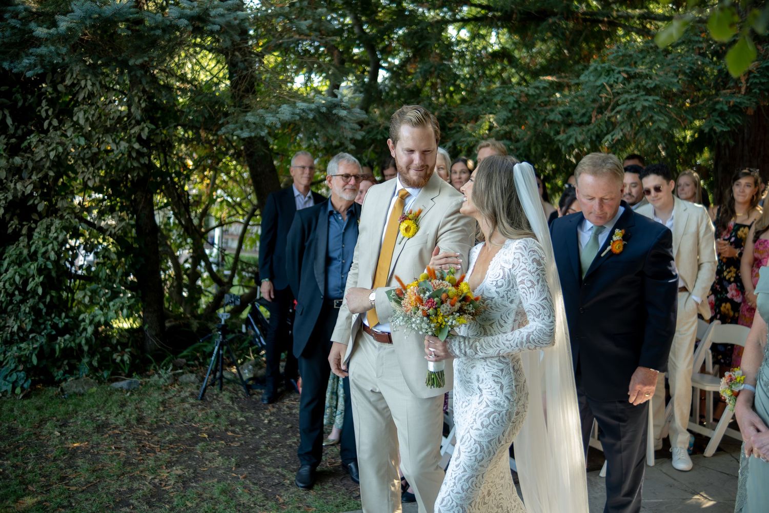 Groom in tan suit walks bride in lace gown down outdoor garden aisle with yellow floral bouquet.