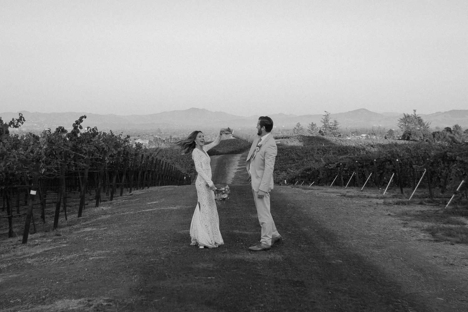 Black and white photo of bride and groom facing each other in vineyard at dusk.
