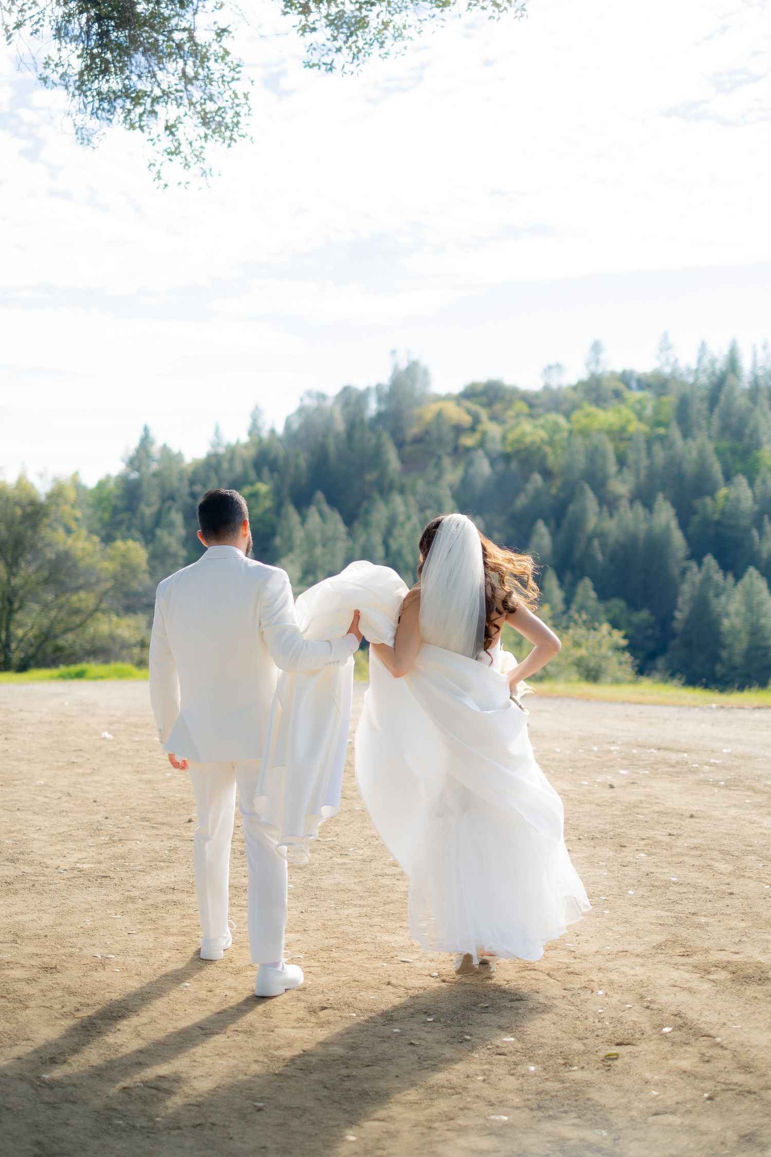Bride and groom in white wedding attire walk hand-in-hand toward lush green forest backdrop on sunny day.