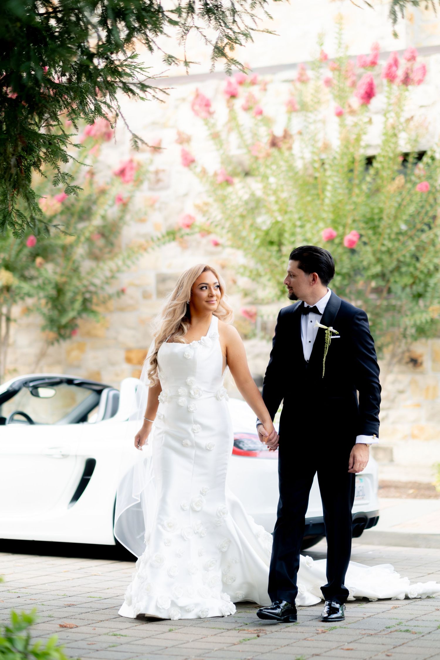 Bride and groom holding hands in front of a white sports car with pink flowering trees in the background.