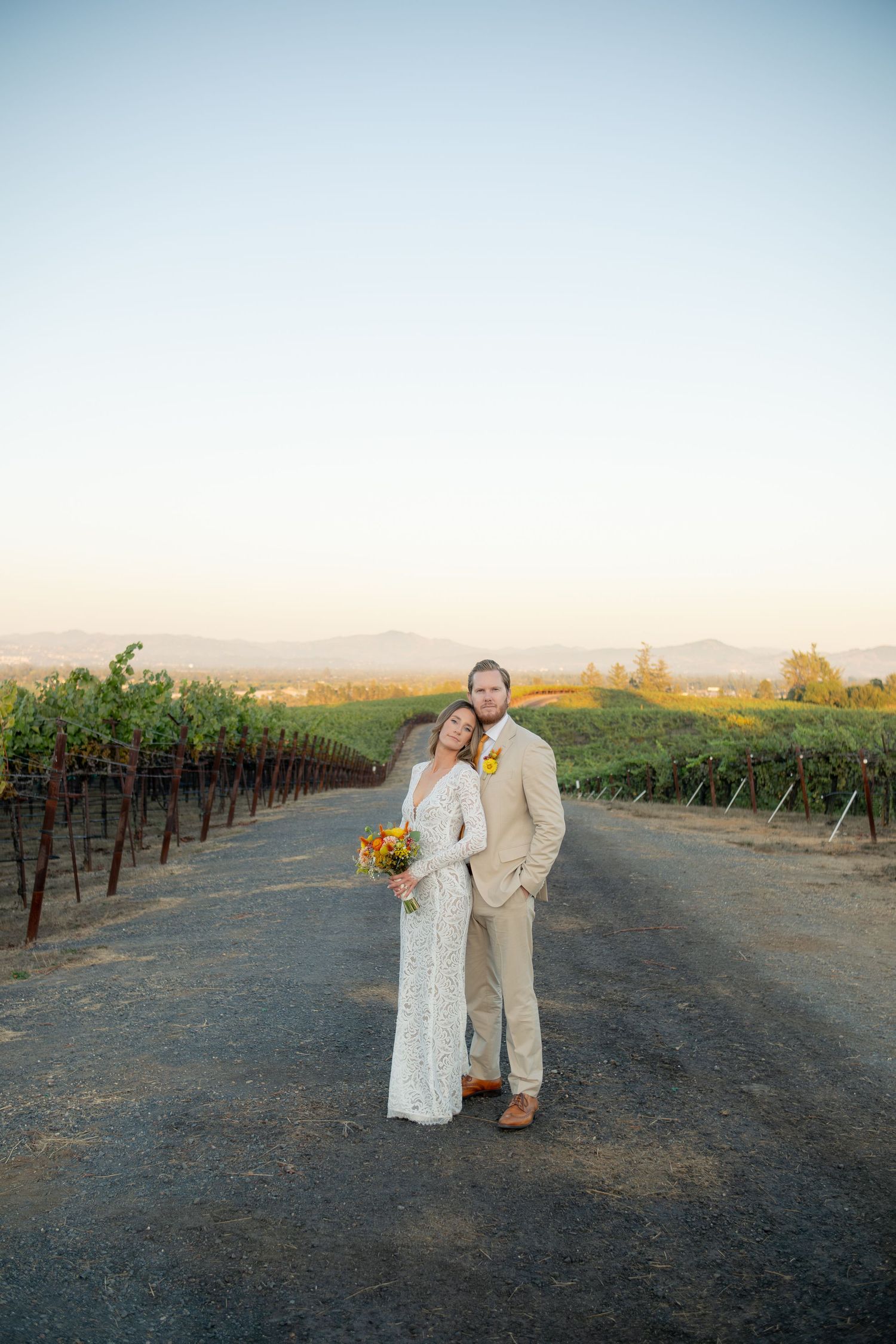 Bride and groom pose at sunset in vineyard rows with mountain backdrop.