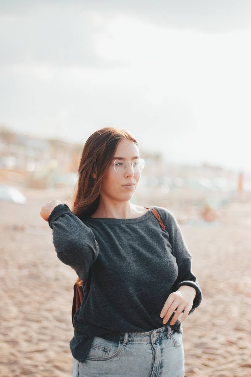 Young woman with long red hair and glasses poses on a sandy beach wearing a gray sweater and jeans.