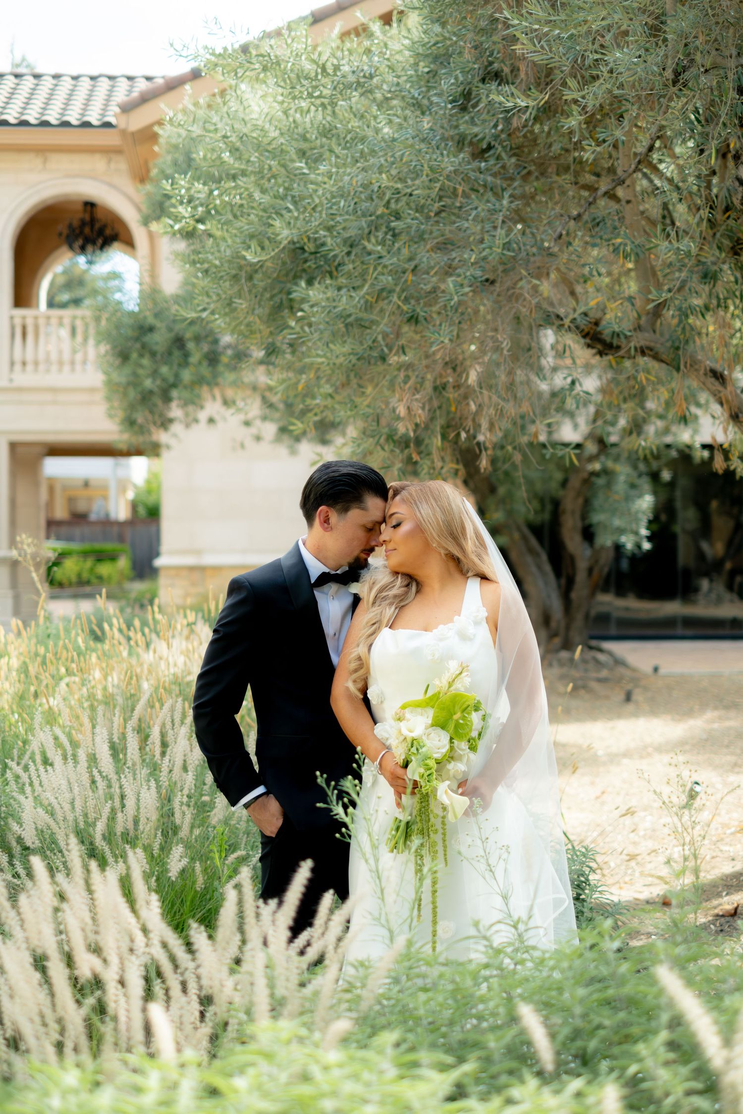Bride and groom share intimate moment in lush garden near Mediterranean-style villa.