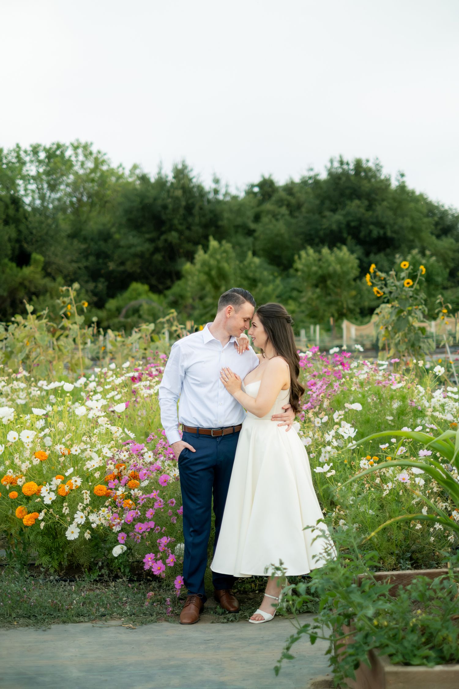 Couple embracing in a colorful wildflower garden, woman in white dress and man in blue pants and white shirt.