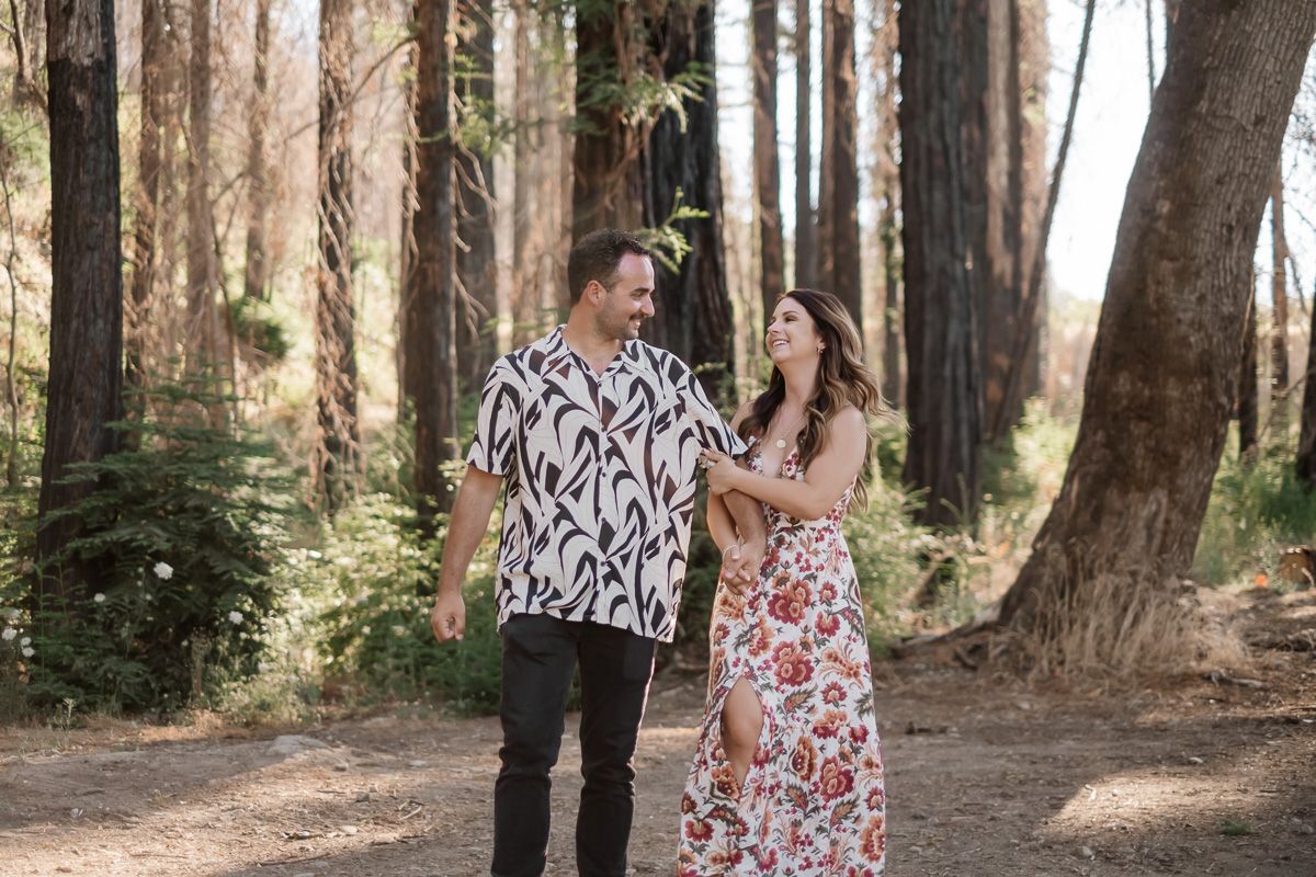 Couple walking hand in hand through a forest path, man in floral shirt and woman in floral dress, smiling at each other.
