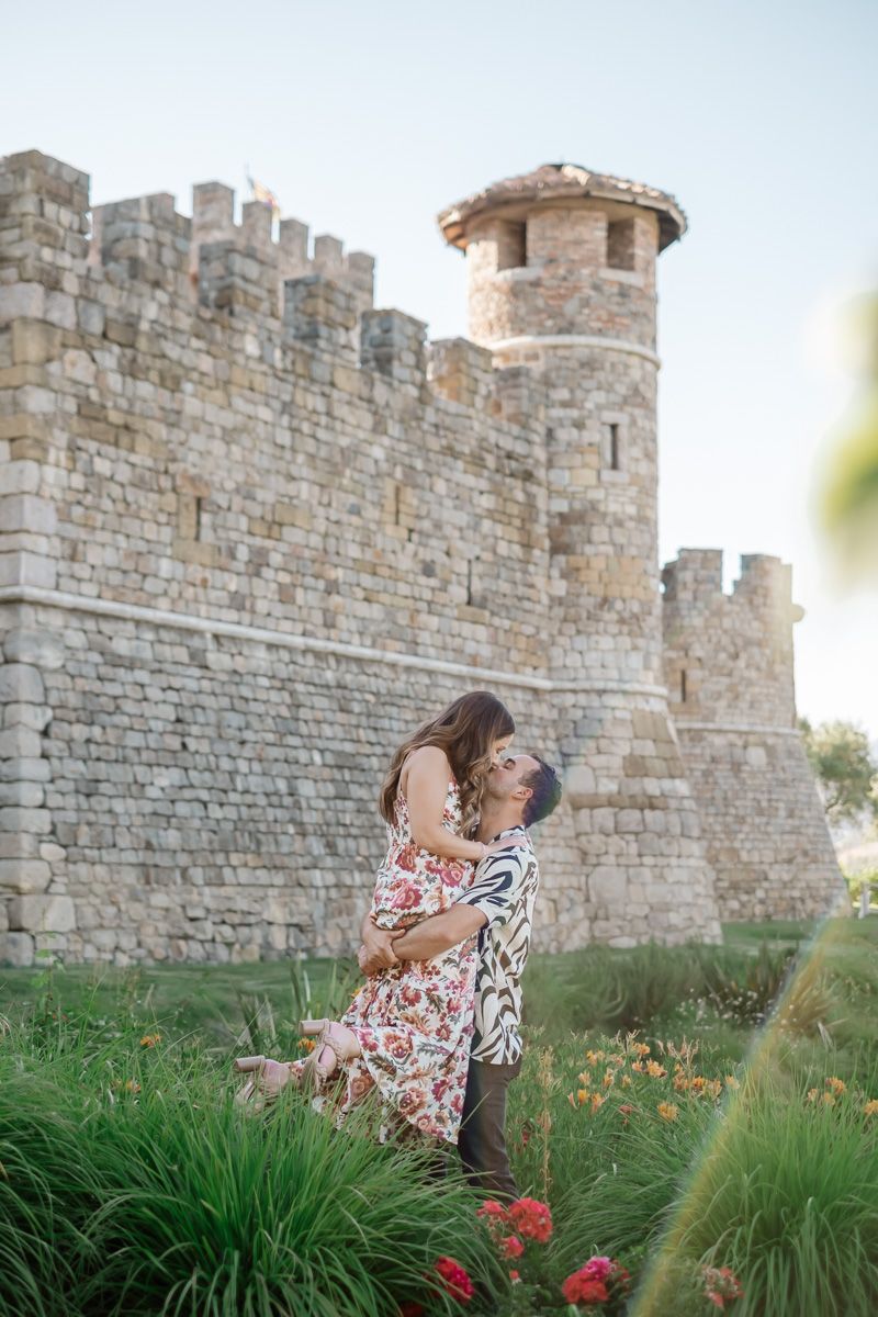 Couple embracing in front of a medieval stone castle surrounded by colorful flowers.