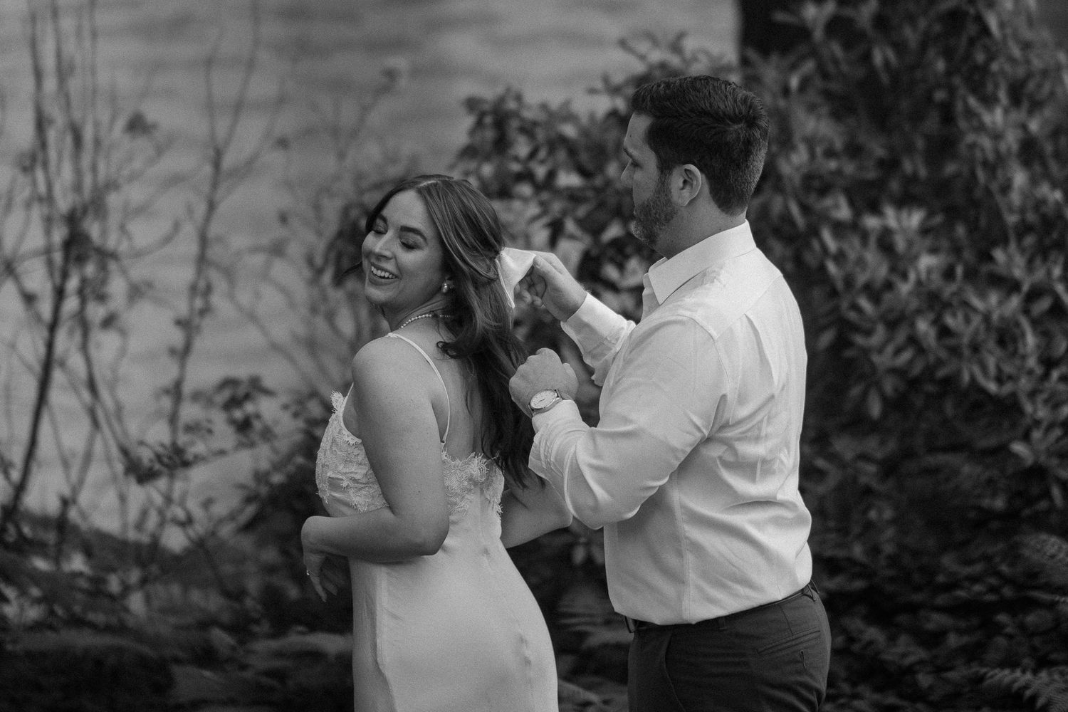Black and white photo of a groom tenderly brushing hair from his smiling bride's face outdoors.