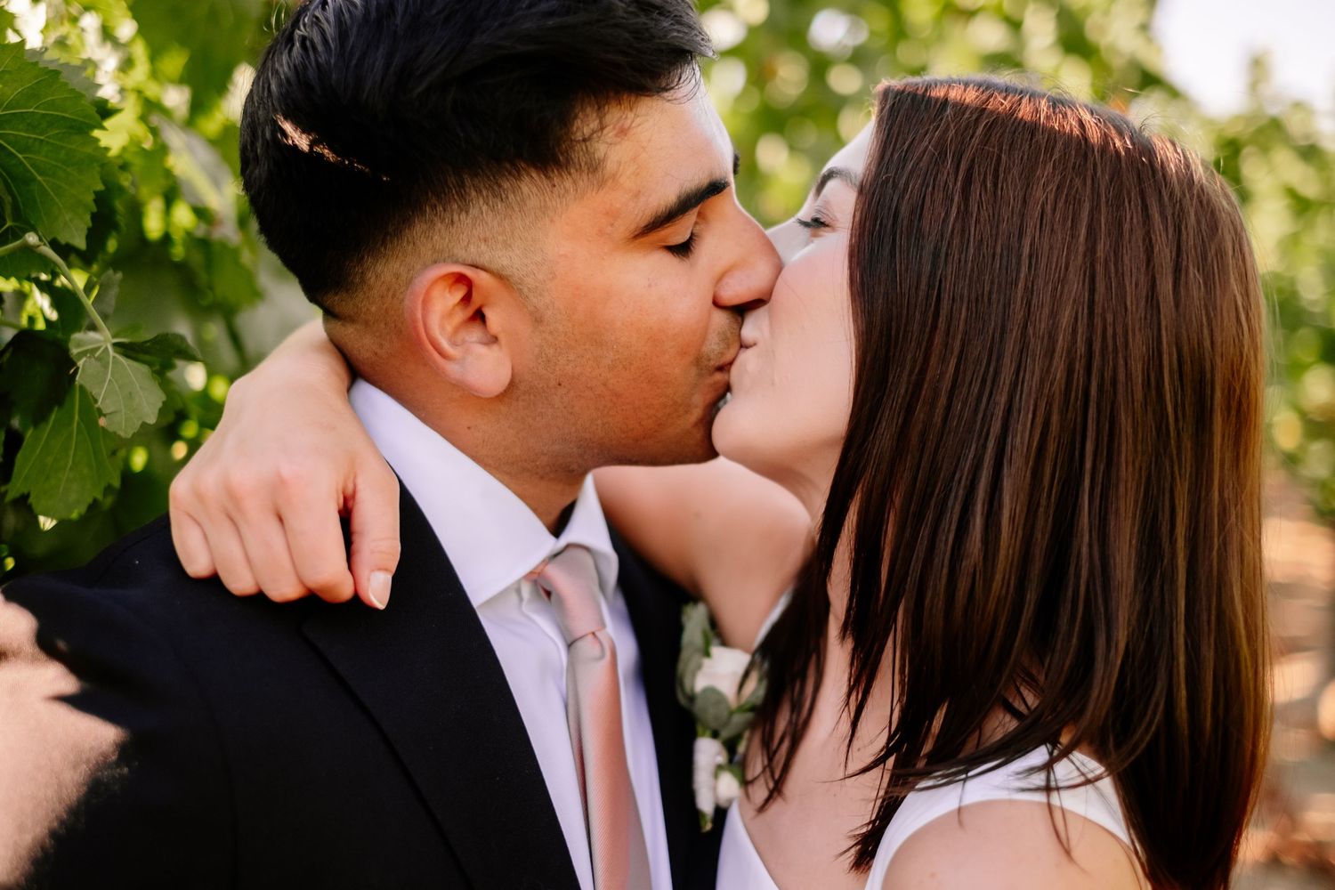 Couple sharing a romantic kiss outdoors in a vineyard, groom in black suit with pink tie, bride in white dress.