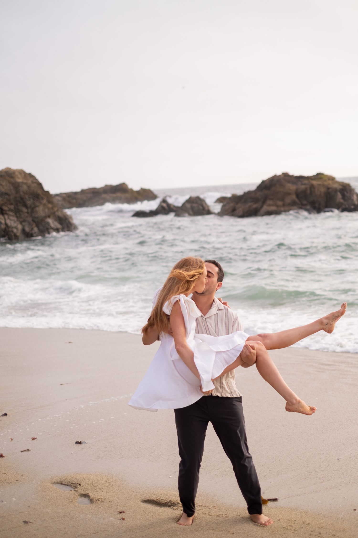 Man carrying woman in his arms on a sandy beach with rocky ocean waves in the background.