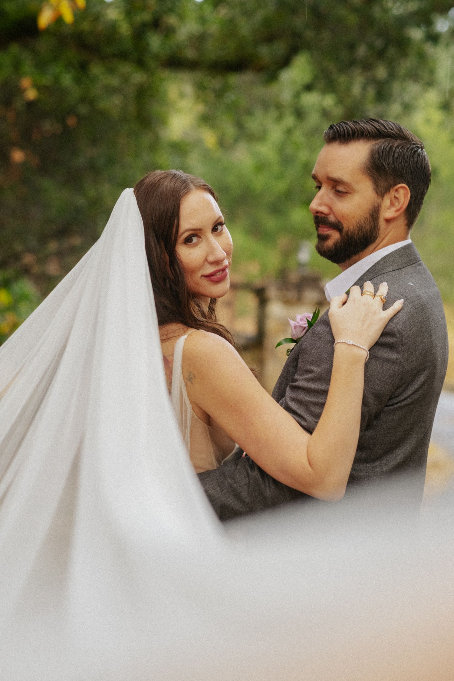 Bride in veil smiling at camera while embracing bearded groom in gray suit outdoors.