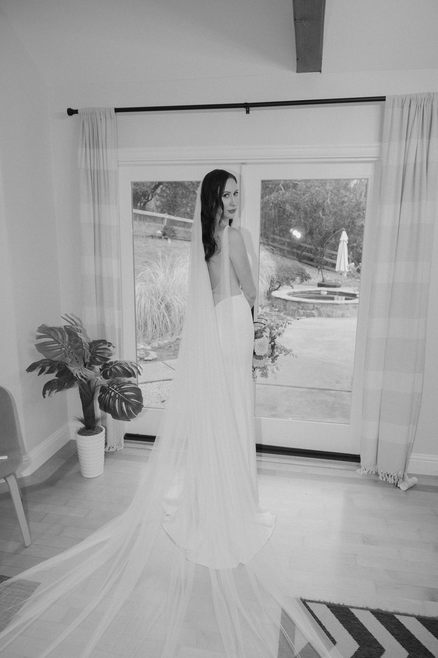 Bride in backless white wedding gown with long veil stands by large window overlooking garden in black and white photo.