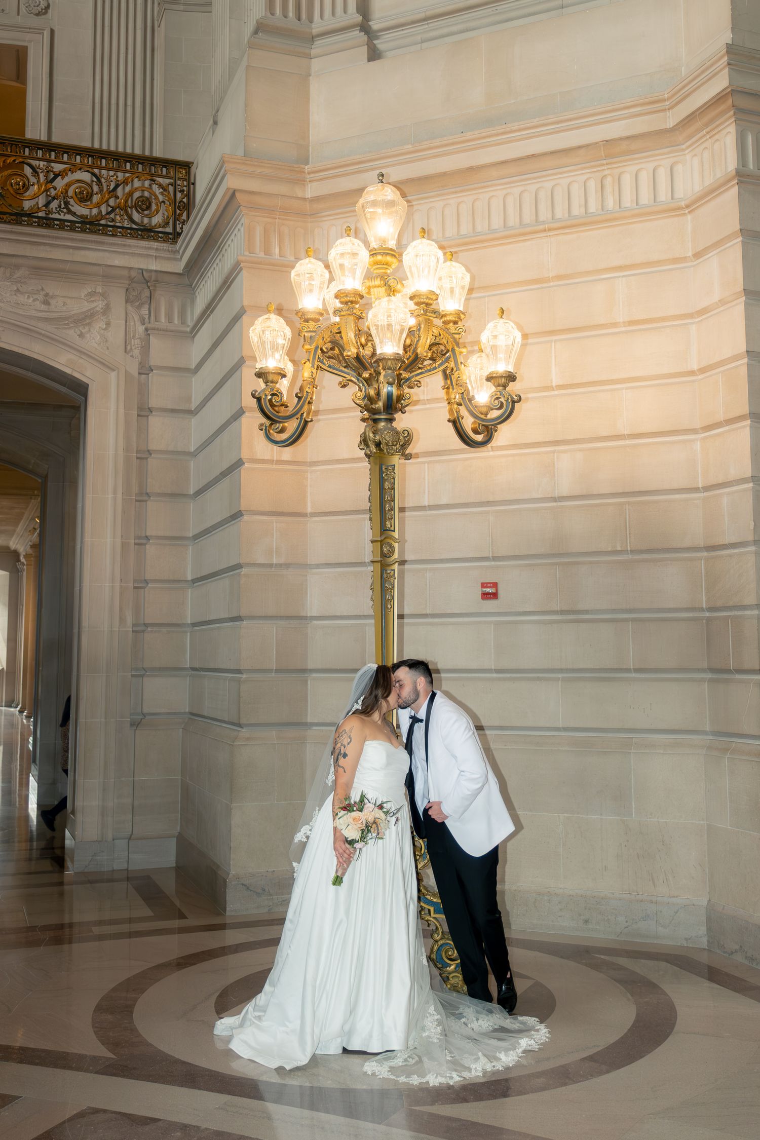 Bride and groom kissing under ornate gold candelabra lamp in elegant marble building interior.
