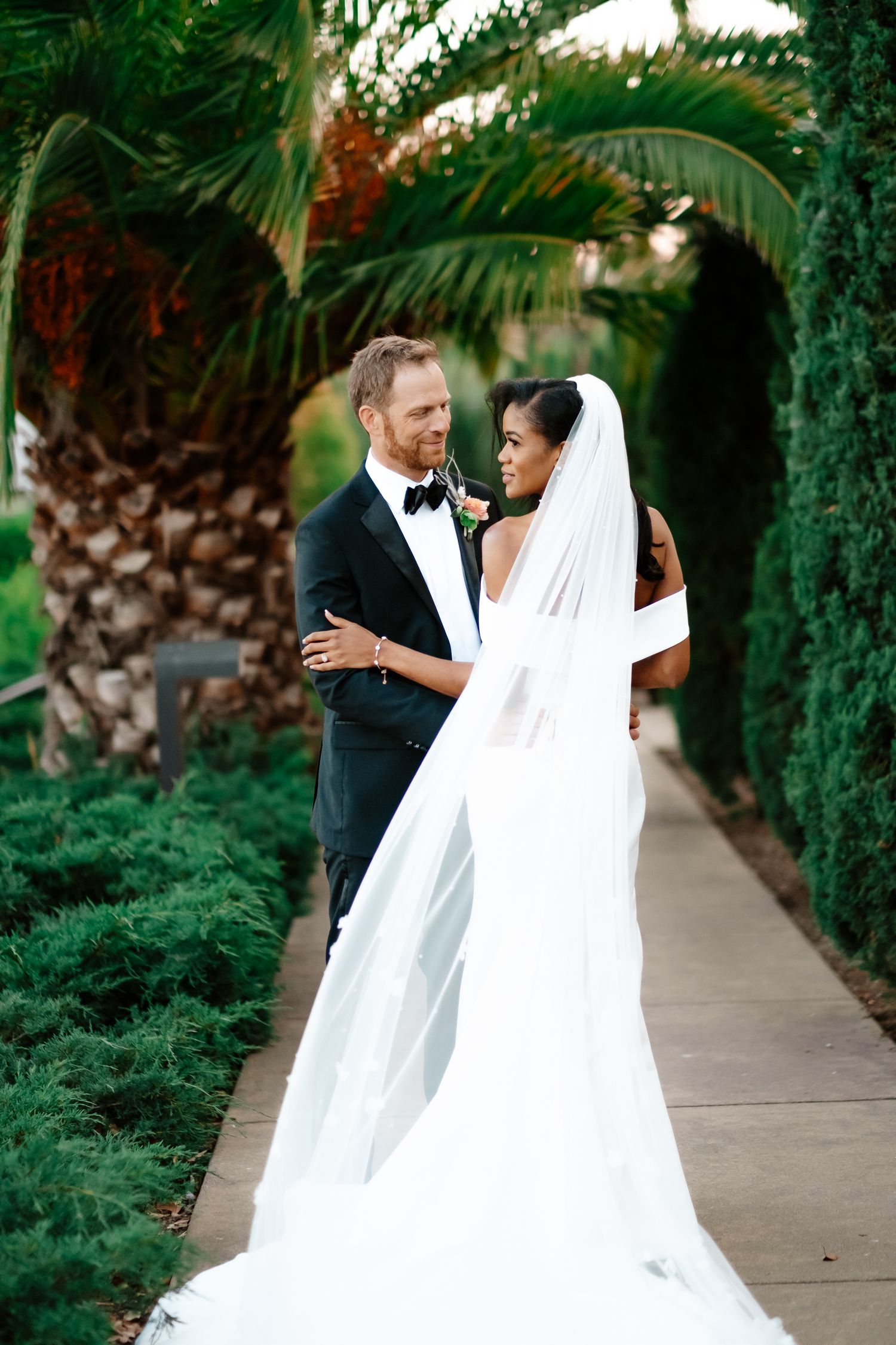 Bride in white gown with long veil and groom in black tuxedo embracing on garden pathway with palms.