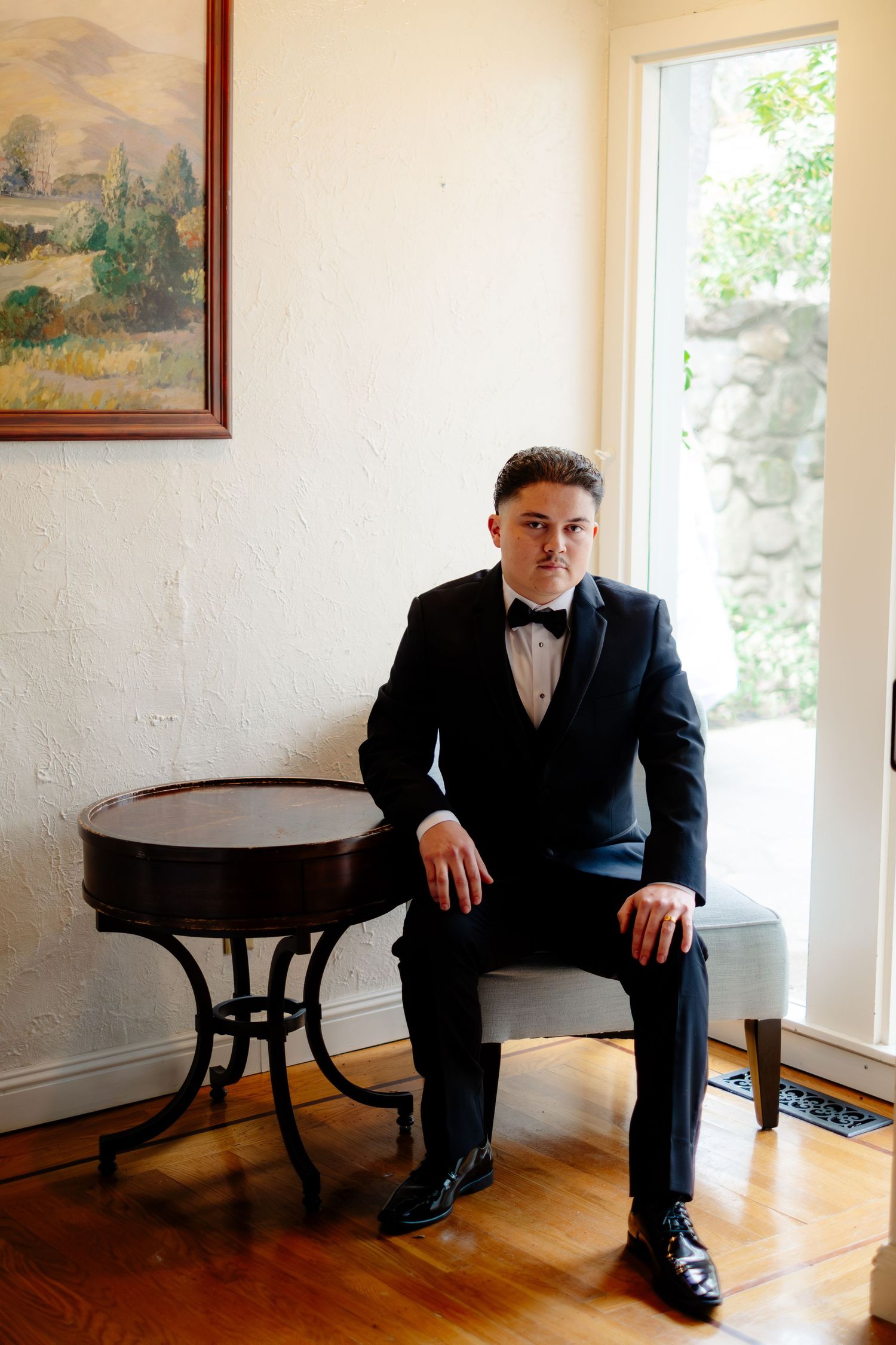 A groom in a black tuxedo and bow tie sits poised on a bench beside a round wooden table indoors.