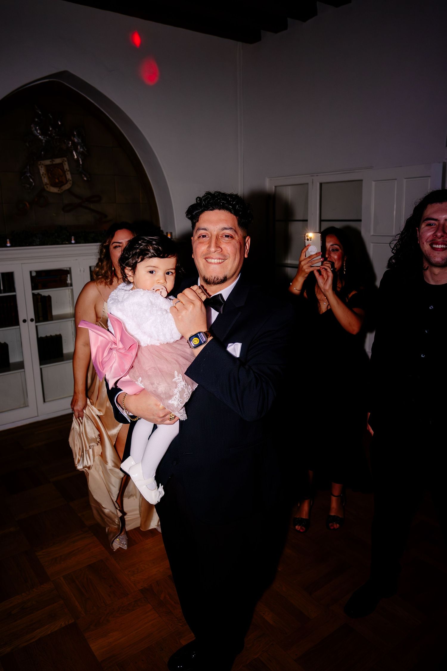 Groom in tuxedo holds young girl in pink dress on wedding reception dance floor.