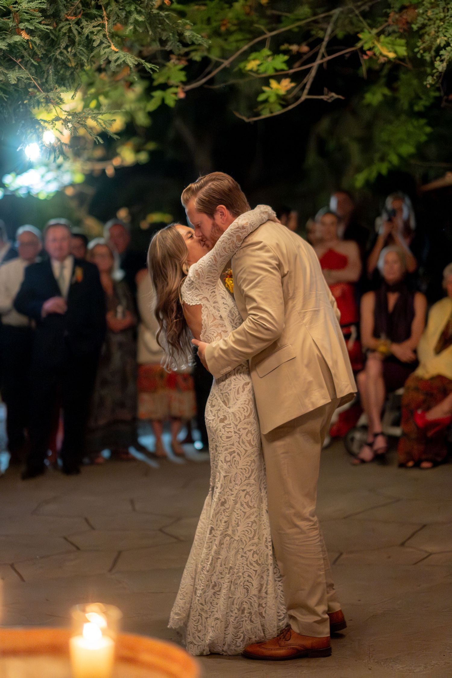 Bride and groom share romantic first dance kiss under trees with candles and guests watching.