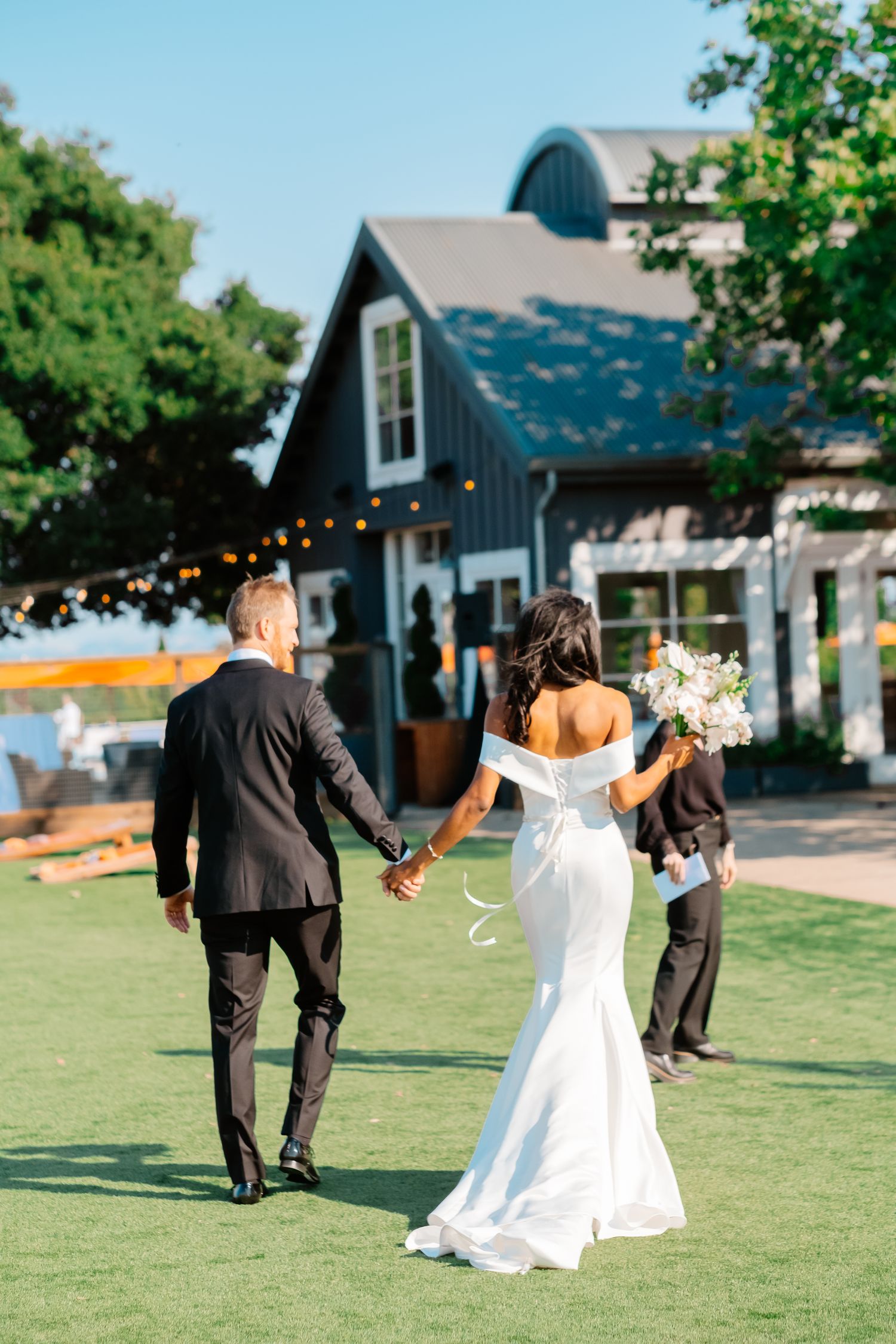 Bride and groom walking hand in hand toward black barn wedding venue on green lawn.