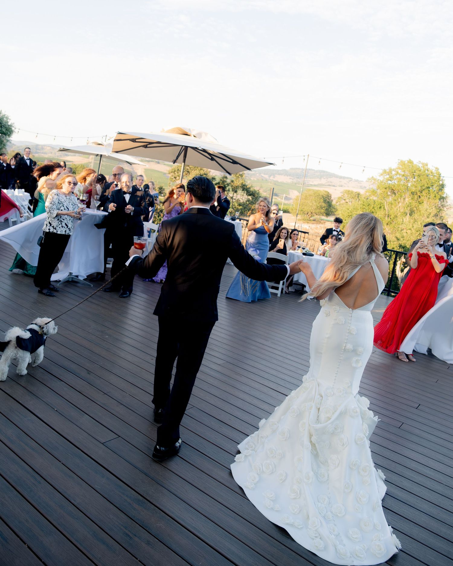 Bride and groom share their first dance on an outdoor deck terrace with wedding guests and scenic views behind them.