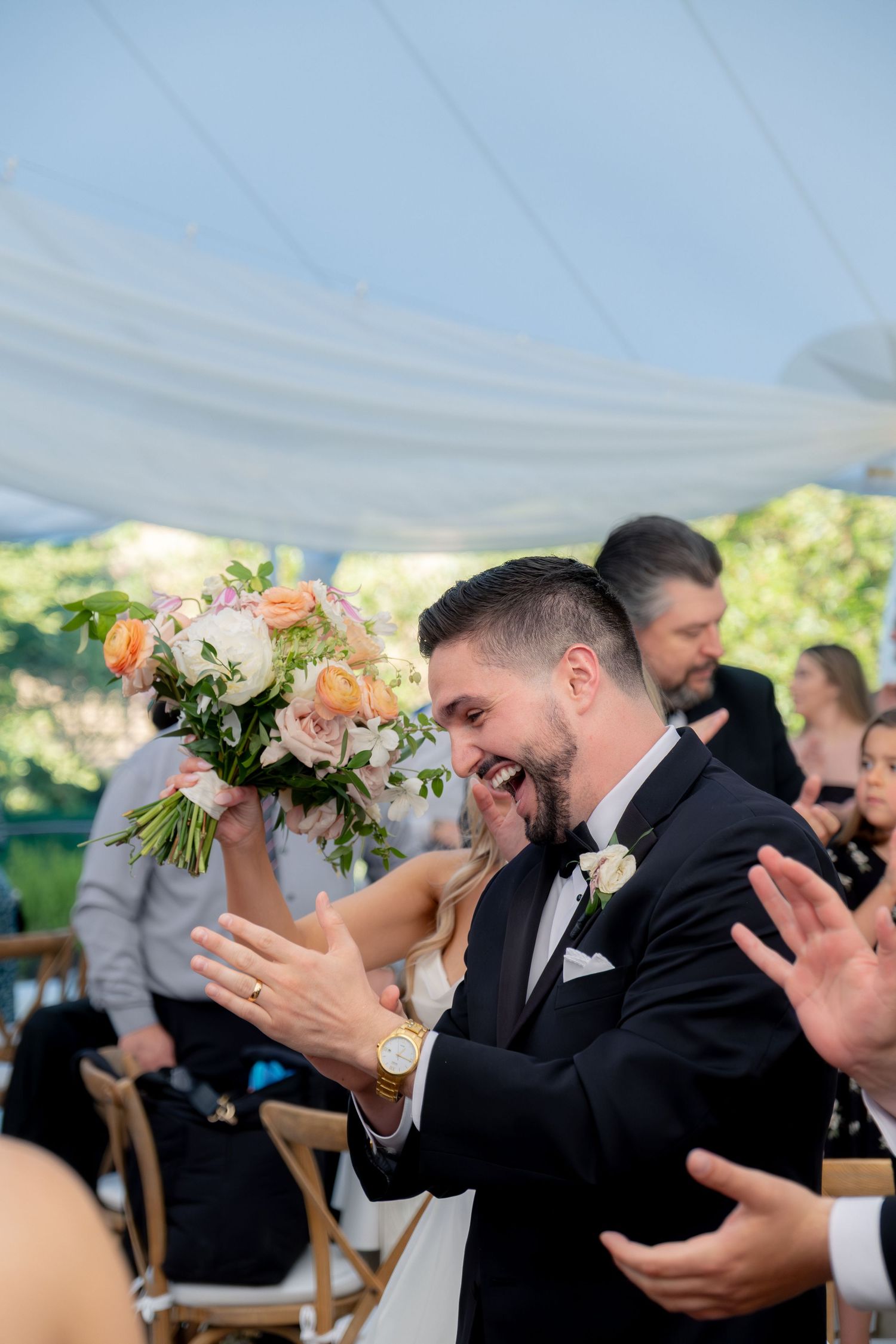 Groom laughs and claps with delight as bride holds bouquet during lively wedding reception entrance.