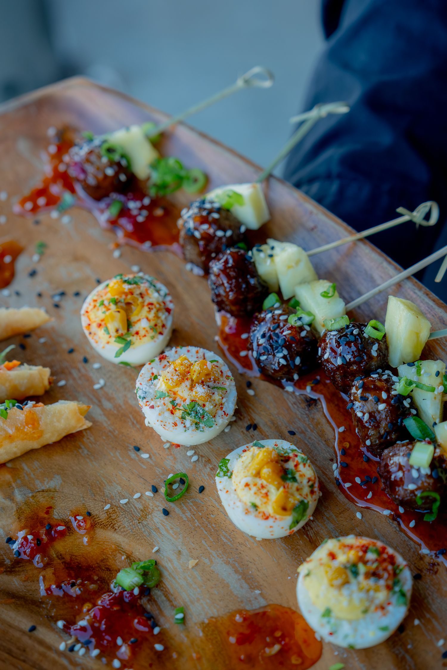 Wooden charcuterie board with deviled eggs, glazed meatball skewers, pineapple bites, and red sauce appetizers.