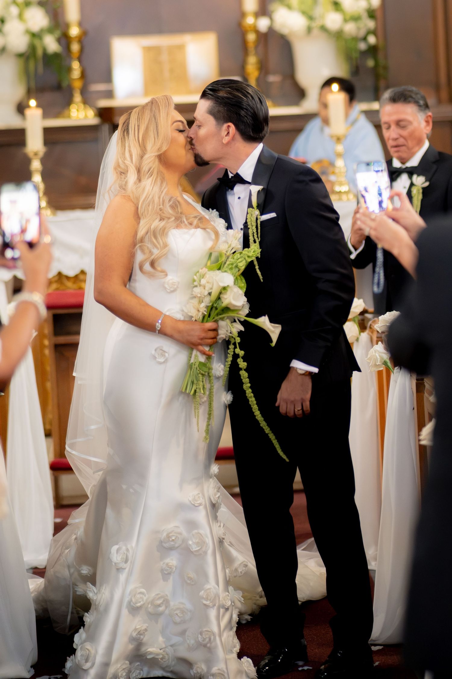 Bride and groom sharing first kiss at church altar while guests capture moment on phones.