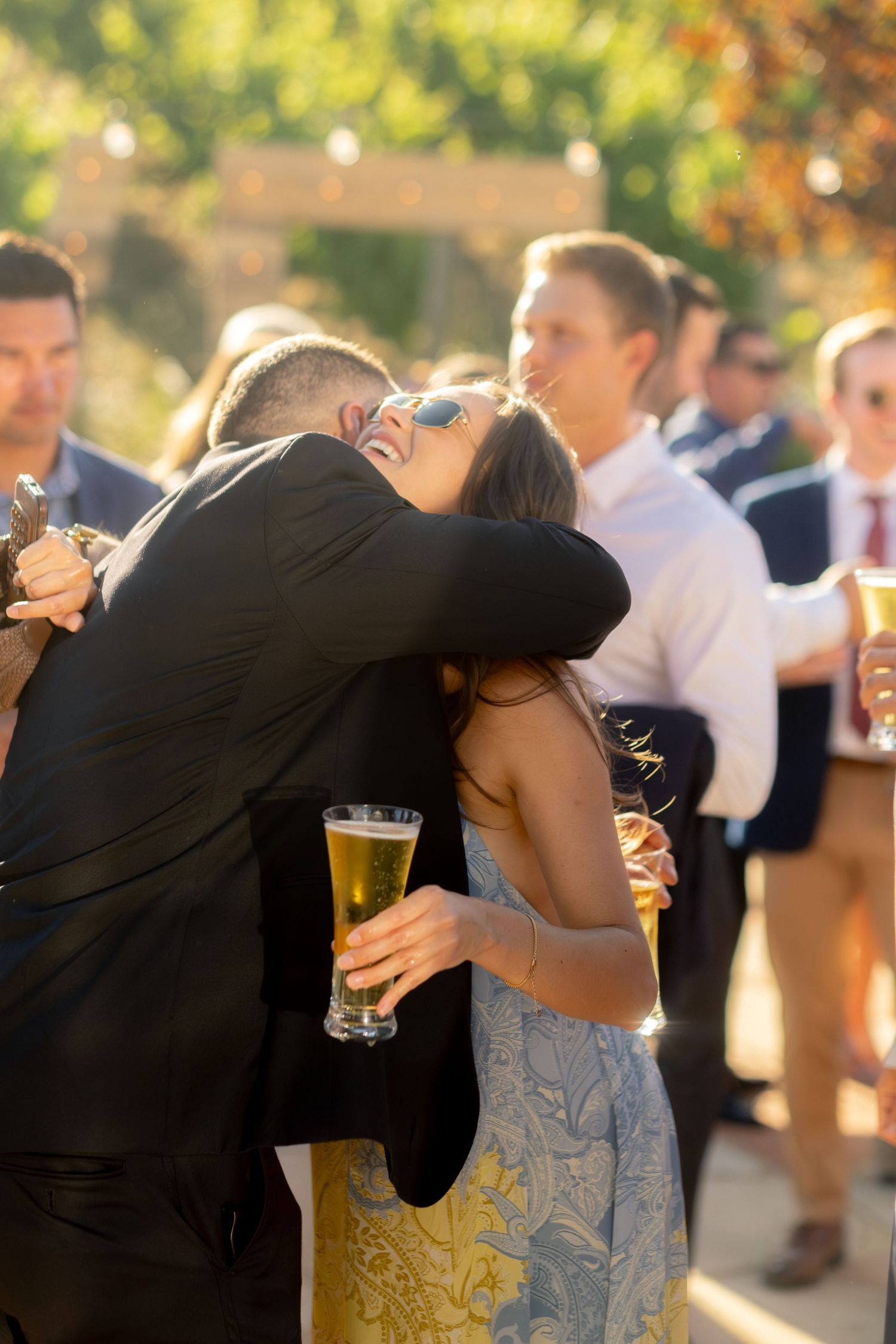Wedding guest in black suit embraces a woman holding a beer glass during outdoor reception.