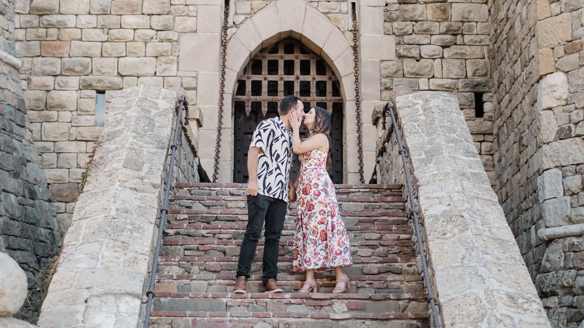 Couple kissing on stone castle steps in a romantic outdoor setting, woman in floral dress, man in patterned shirt.