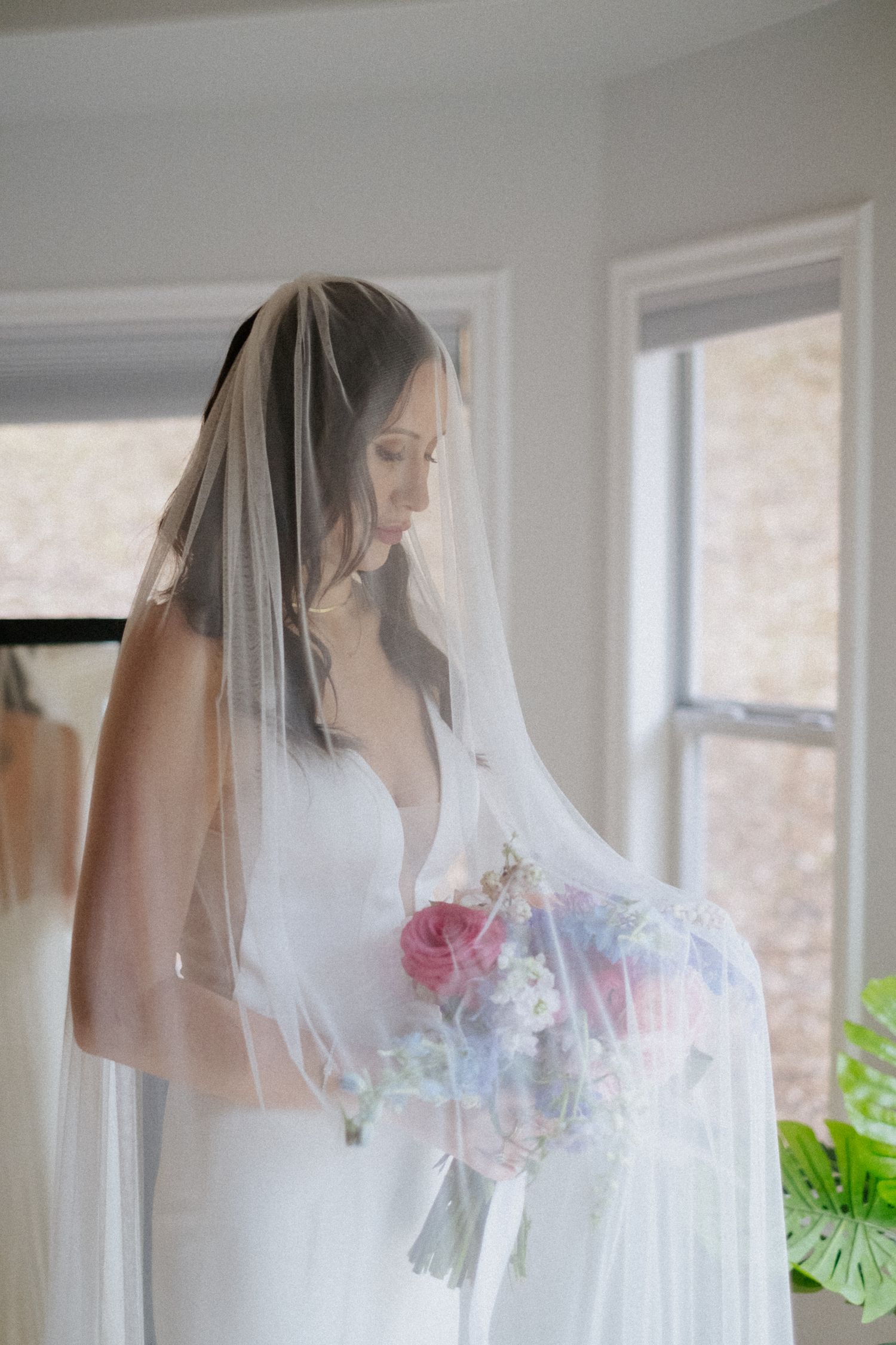 Bride in deep-V gown holds colorful bouquet beneath sheer veil in softly lit bridal suite.