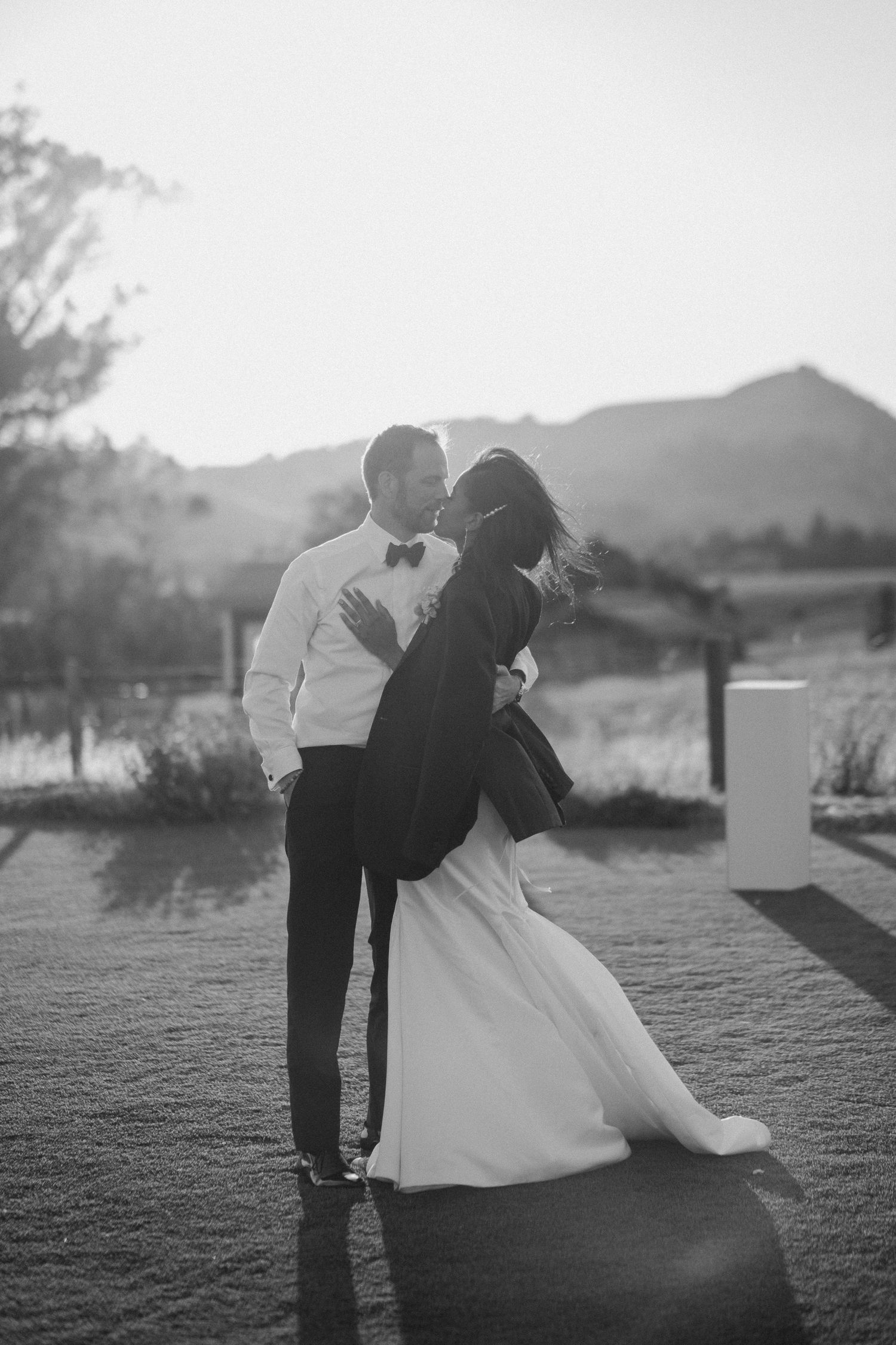 Black and white photo of newlyweds sharing a romantic kiss at golden hour.