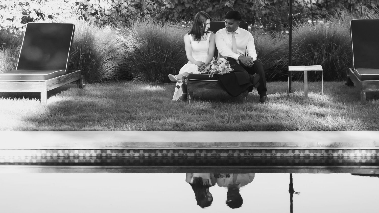Couple sitting together on a bench by a reflective pool in a black and white outdoor engagement photo.