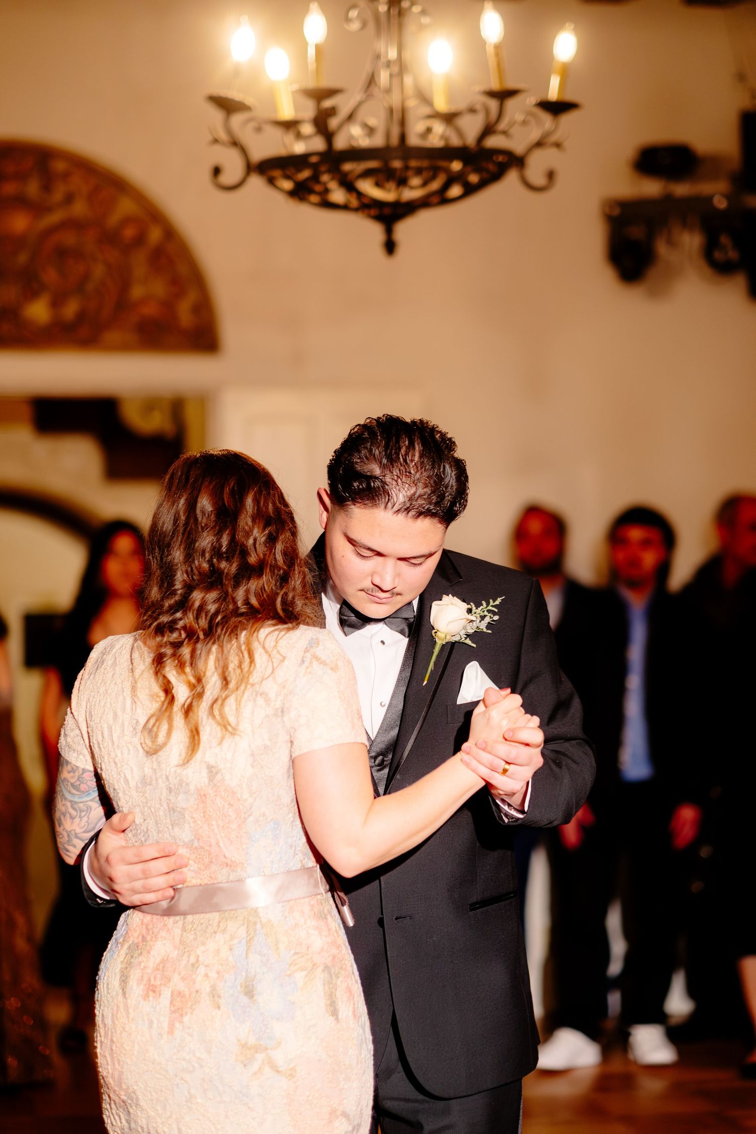 Groom in black tuxedo shares emotional first dance under ornate chandelier at elegant wedding reception.