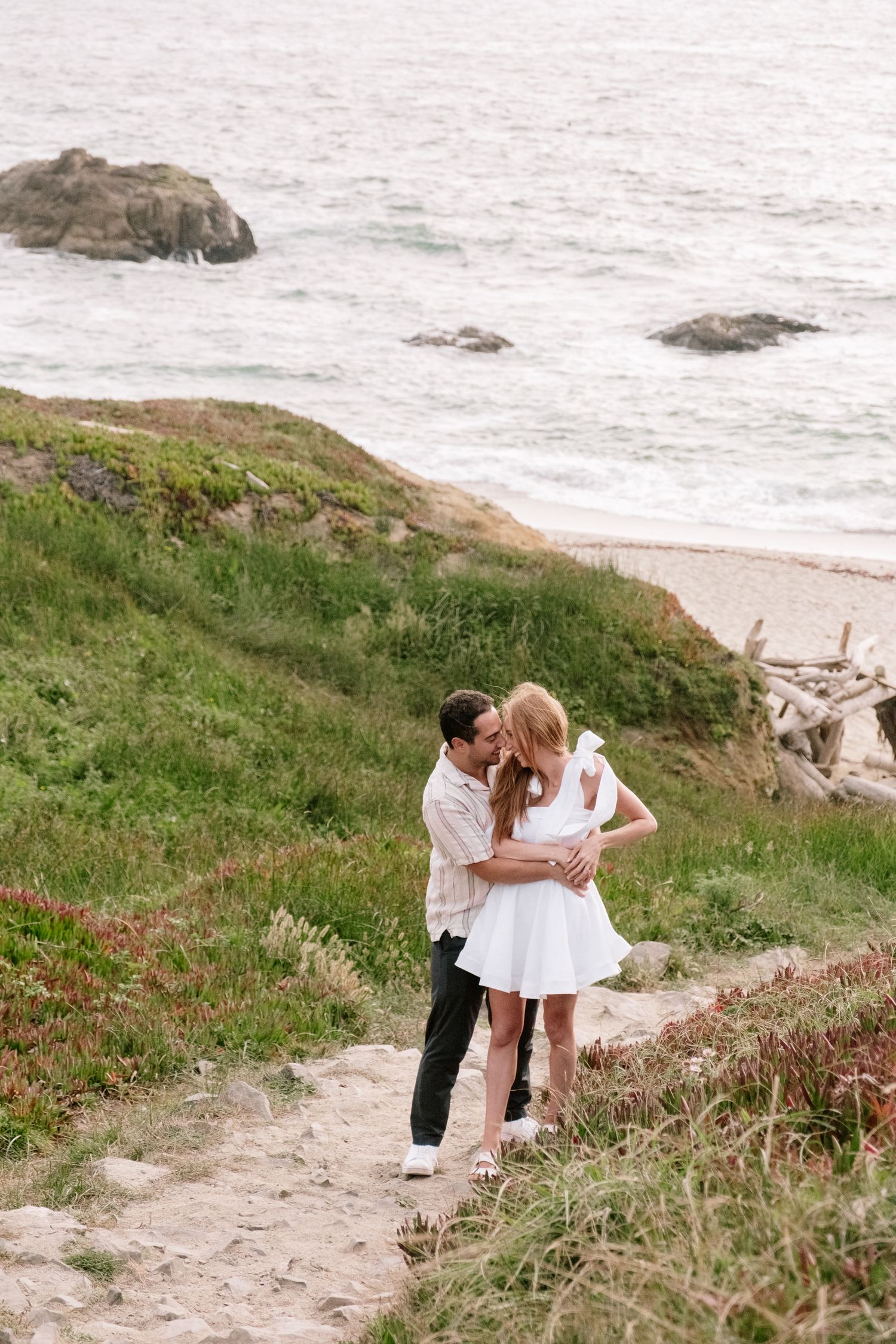 A couple embraces and kisses on a grassy coastal path with the ocean and beach below.
