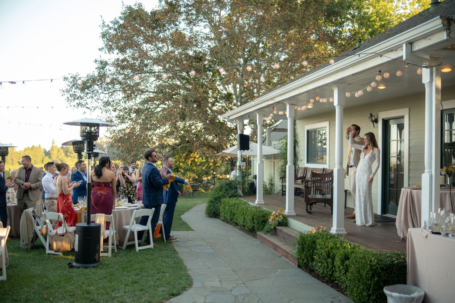 Outdoor wedding reception on a garden pathway beside a white porch venue with string lights and guests mingling at dusk.