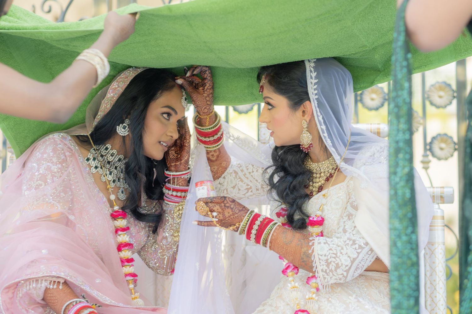Two women in traditional South Asian bridal attire share a moment under a green canopy at a wedding ceremony.