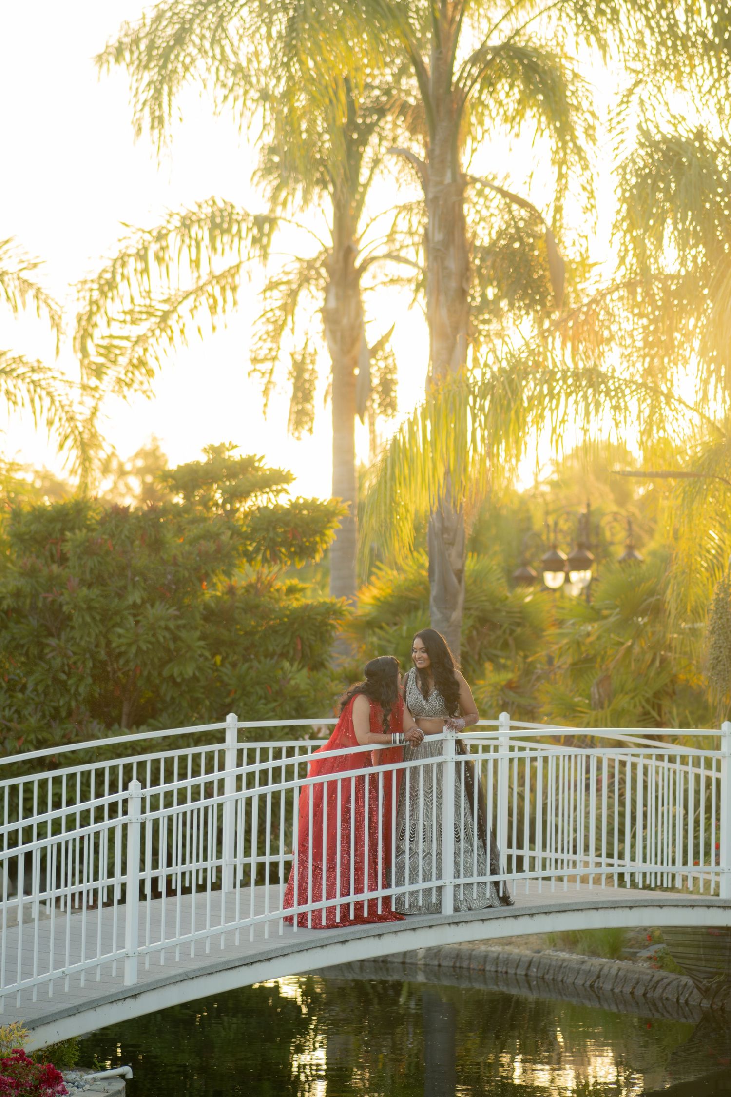 Two women in formal gowns stand together on white arched bridge at golden sunset among palm trees.