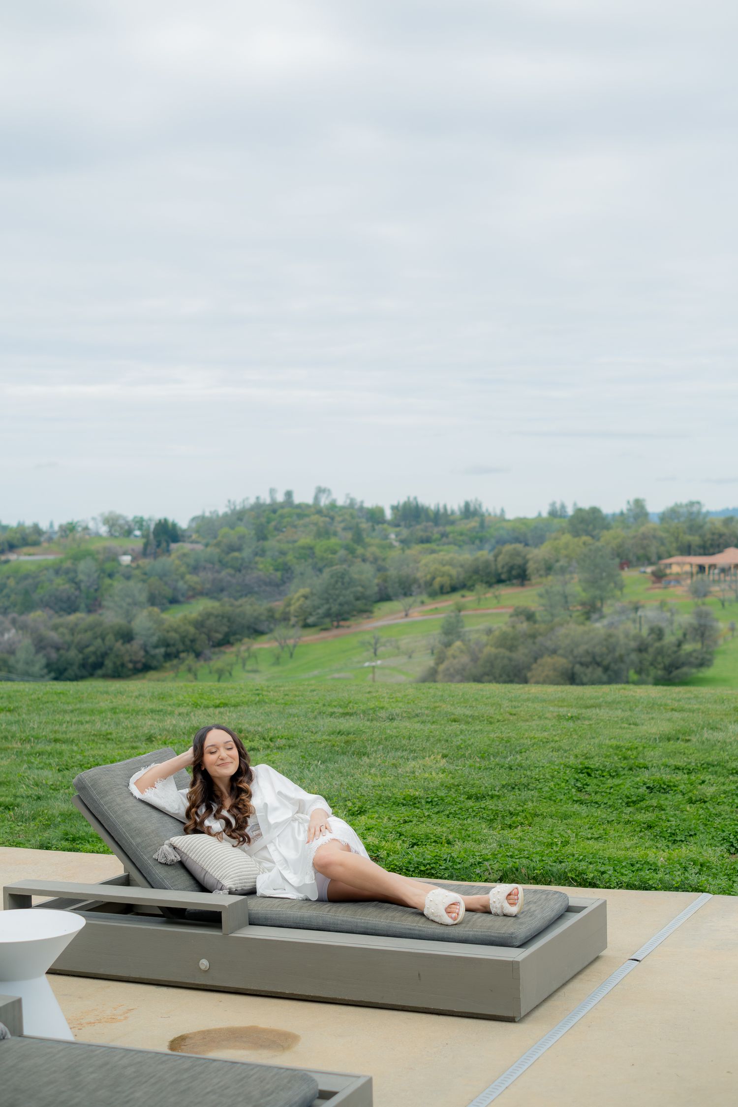 Woman in white dress relaxing on outdoor chaise lounge with lush green hillside landscape in background.