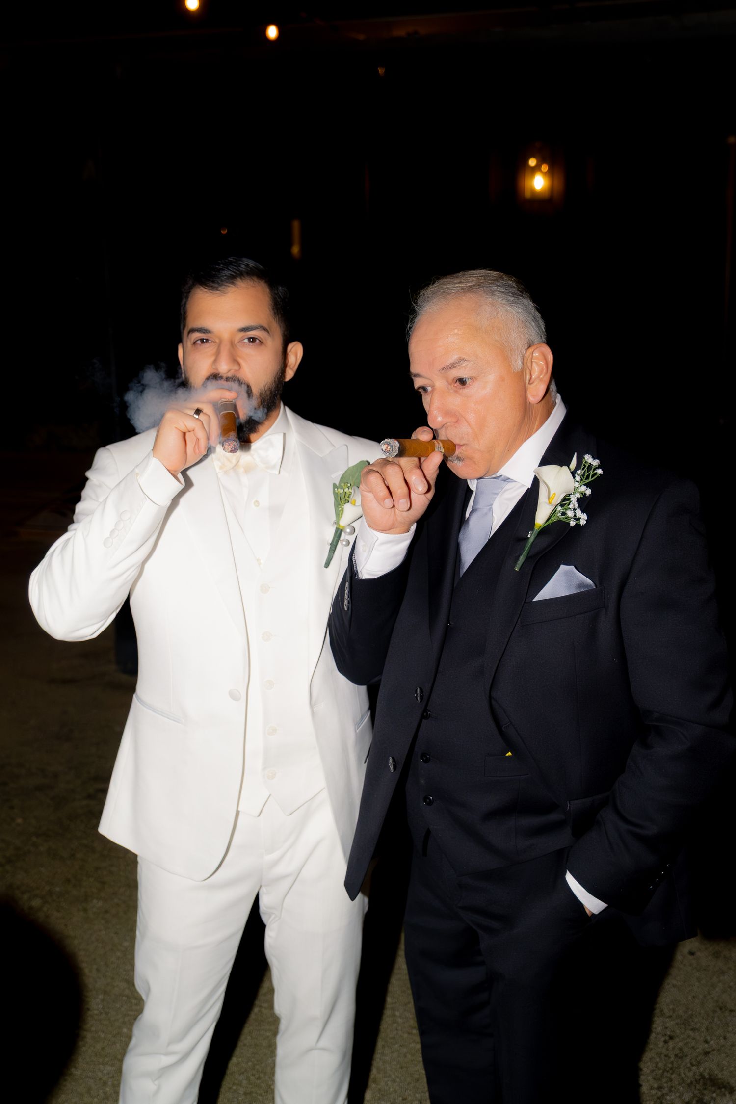 Groom in white suit and older gentleman in navy tuxedo smoke cigars together at night.