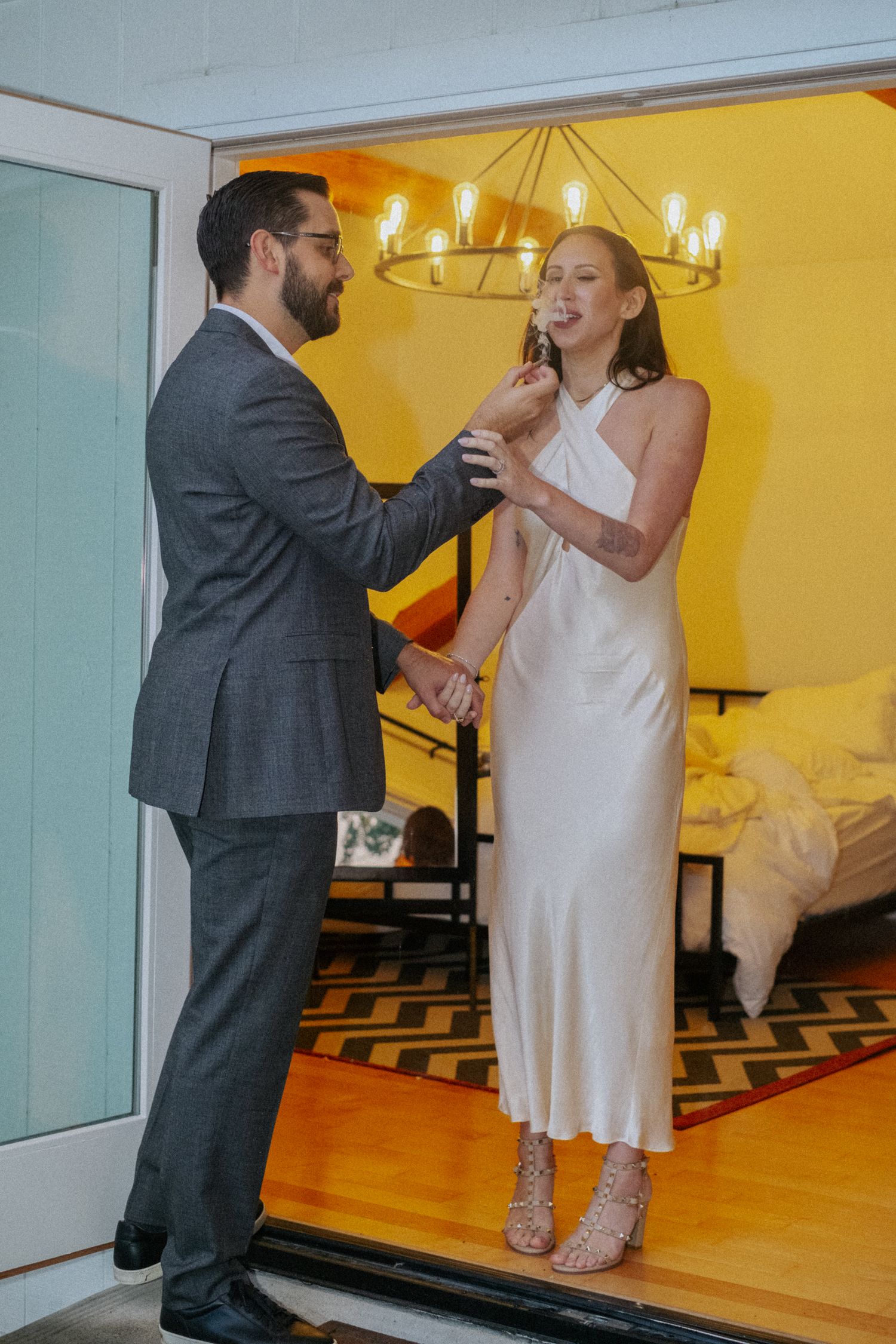 Couple sharing a sweet moment during wedding cake cutting in a stylish room with chandelier.
