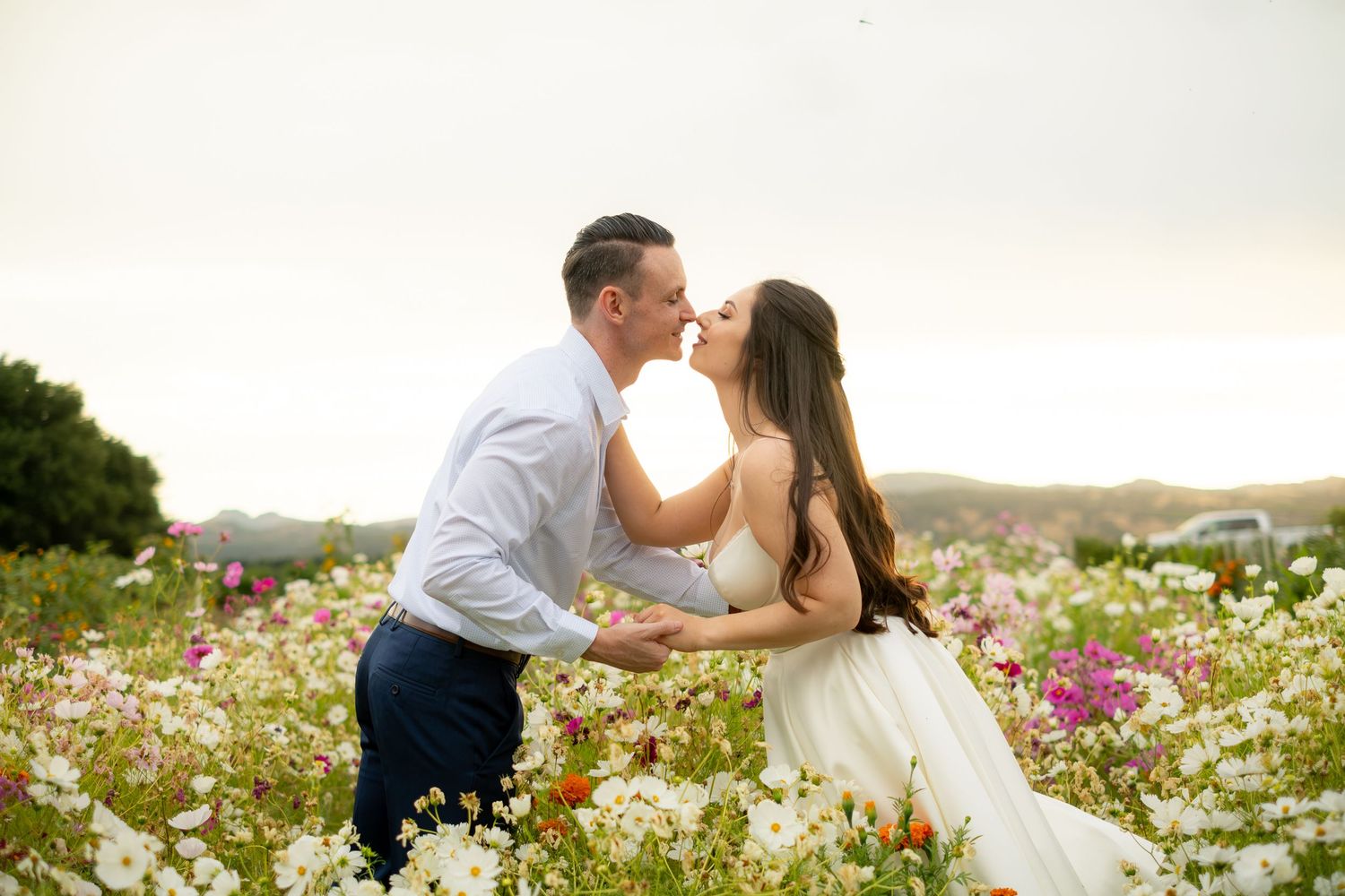 Couple sharing a romantic kiss in a vibrant flower field with white and pink blooms at sunset.