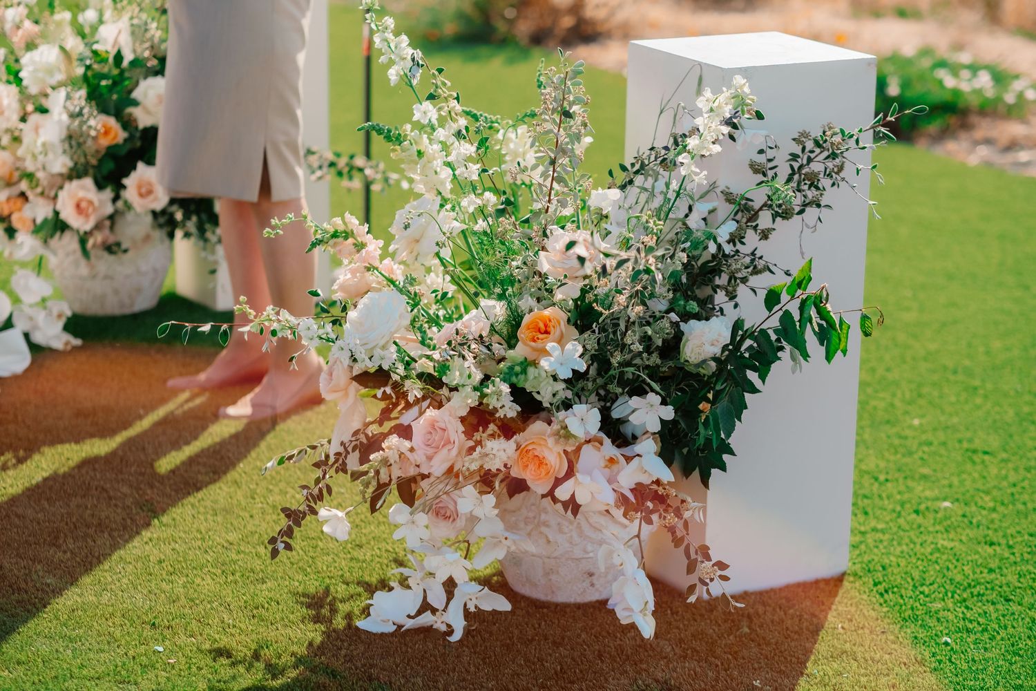 Elegant wedding floral arrangement with white and peach blooms in a white vase on green grass.
