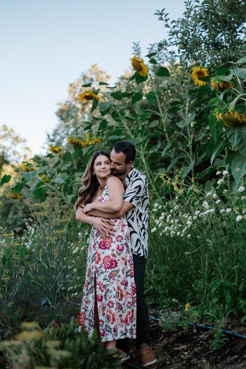 Couple embracing in a sunflower garden, woman in floral dress, man in black and white shirt.