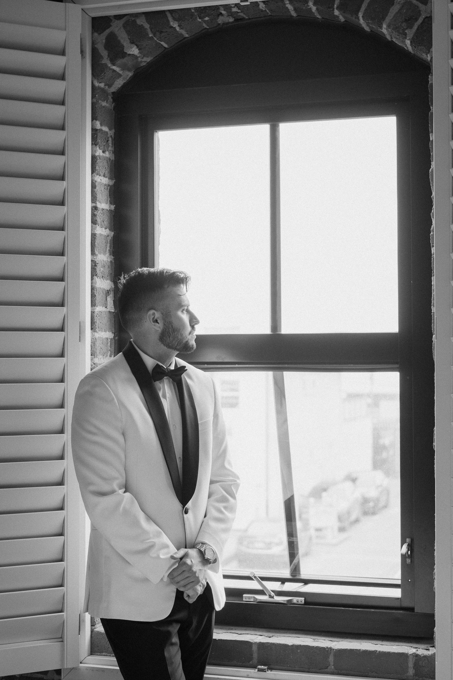 Groom in white tuxedo jacket and bow tie standing by arched window in black and white wedding photo.