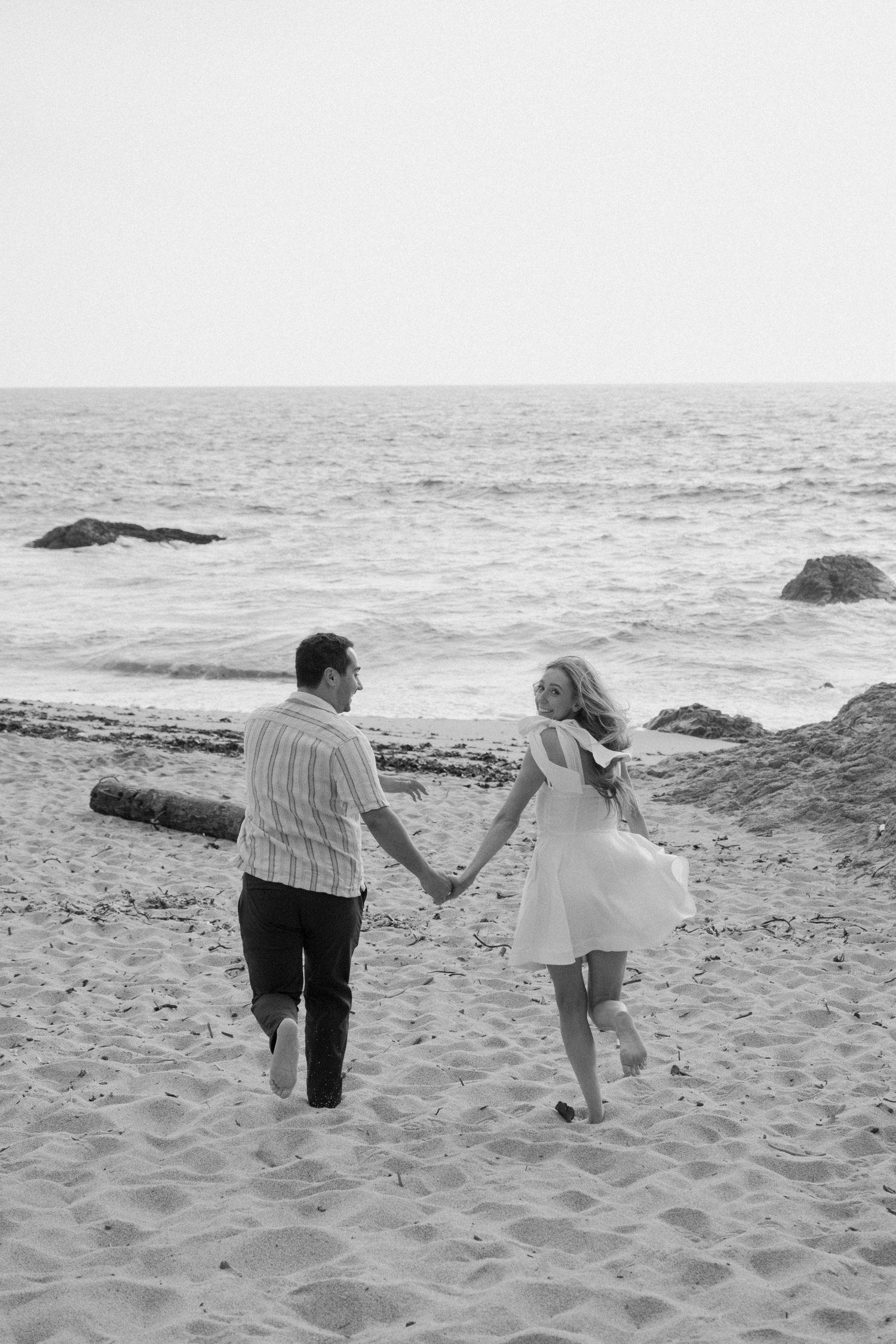 Black and white photo of a couple holding hands walking along a sandy beach toward the ocean.