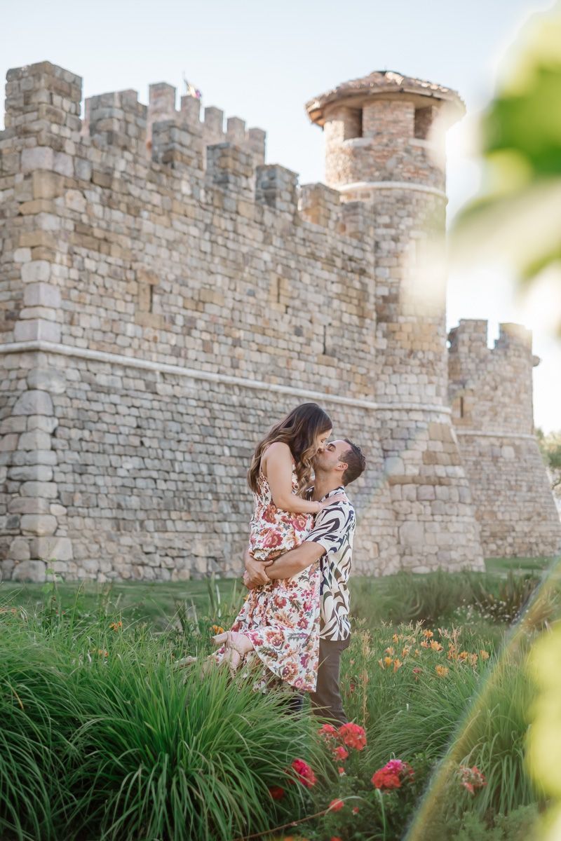 Couple embracing and laughing in front of a rustic stone castle surrounded by vibrant flowers and greenery.